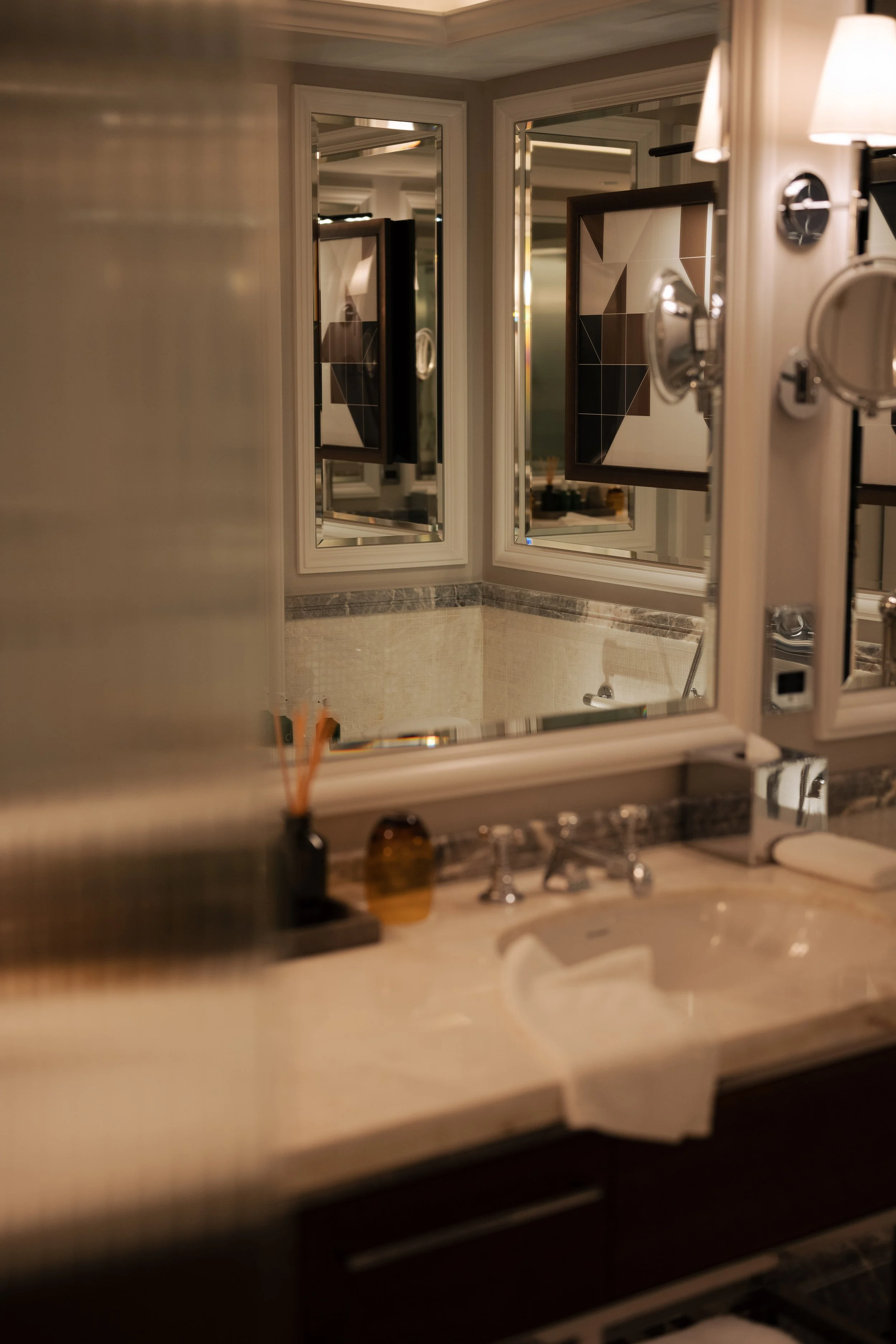 A bathroom with multiple mirrors reflecting a bathtub. The sink countertop has toiletries, a soap dispenser, and a tissue box. Wall-mounted lights illuminate the space.