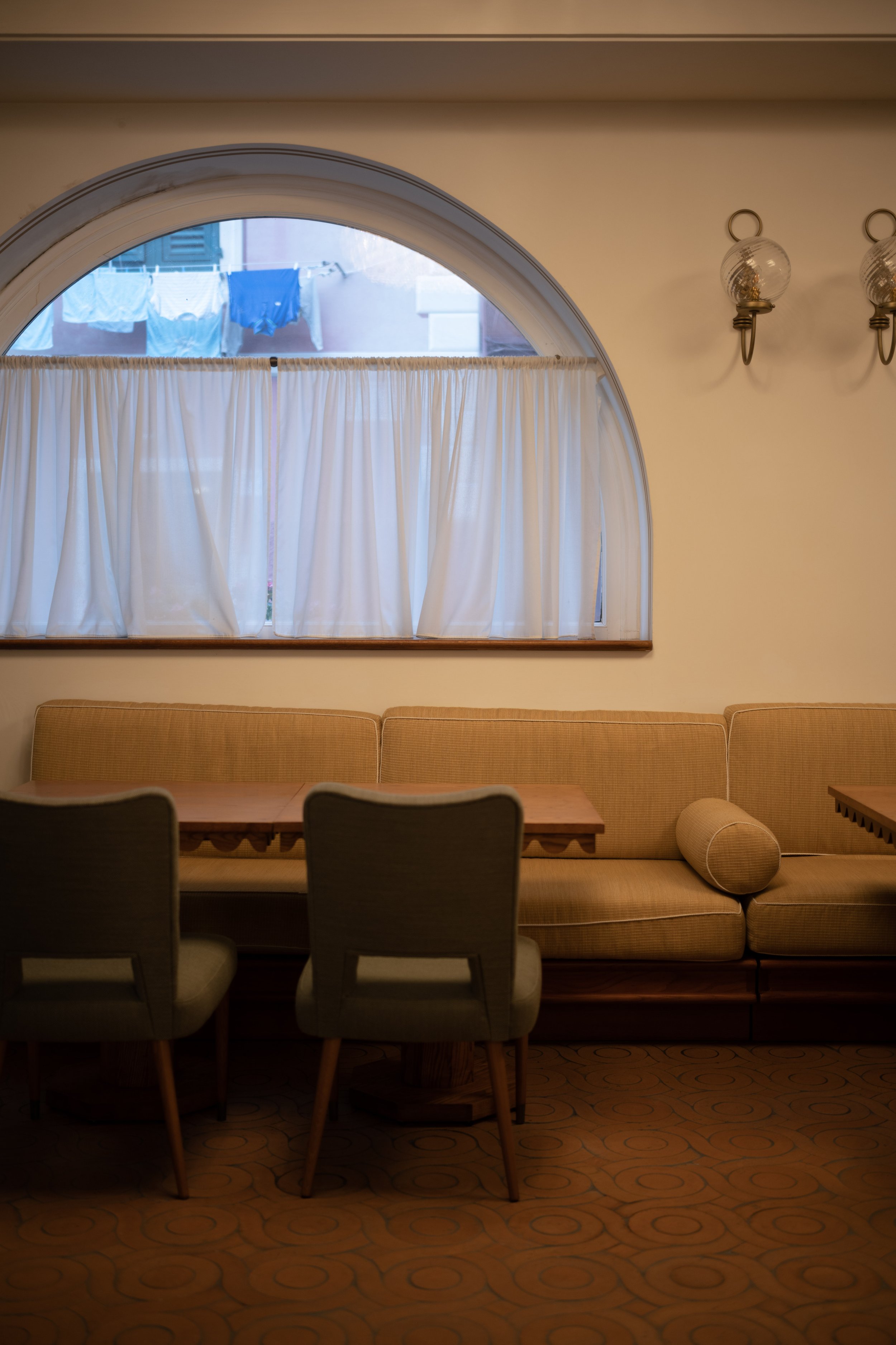 Cozy dining area with beige upholstered chairs, a wooden table, a yellow sofa with a round bolster pillow, and a large window with white curtains, above which laundry is hanging outside.