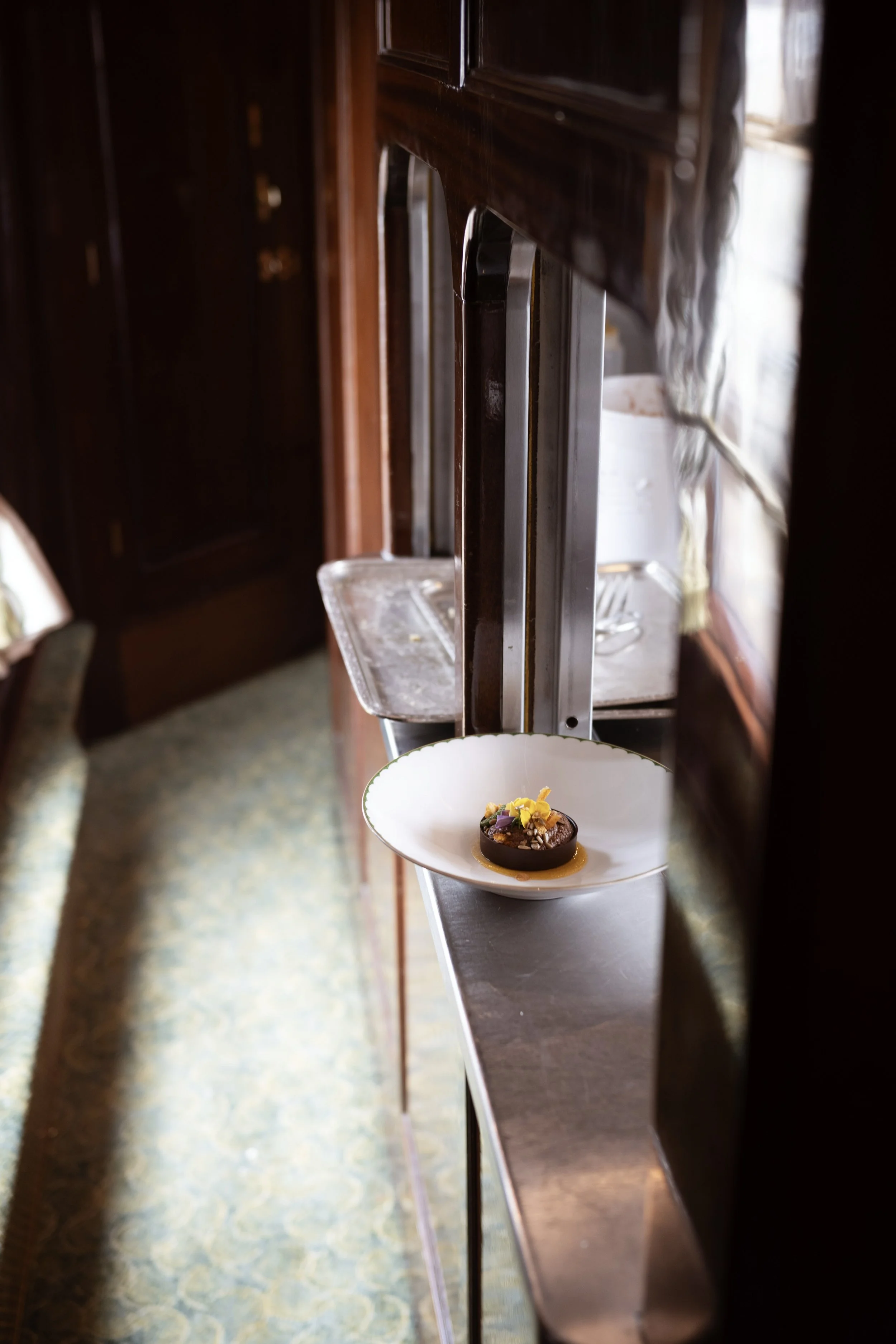 A chocolate dessert garnished with yellow flowers on a white plate on a metal shelf next to dark wooden cabinets.