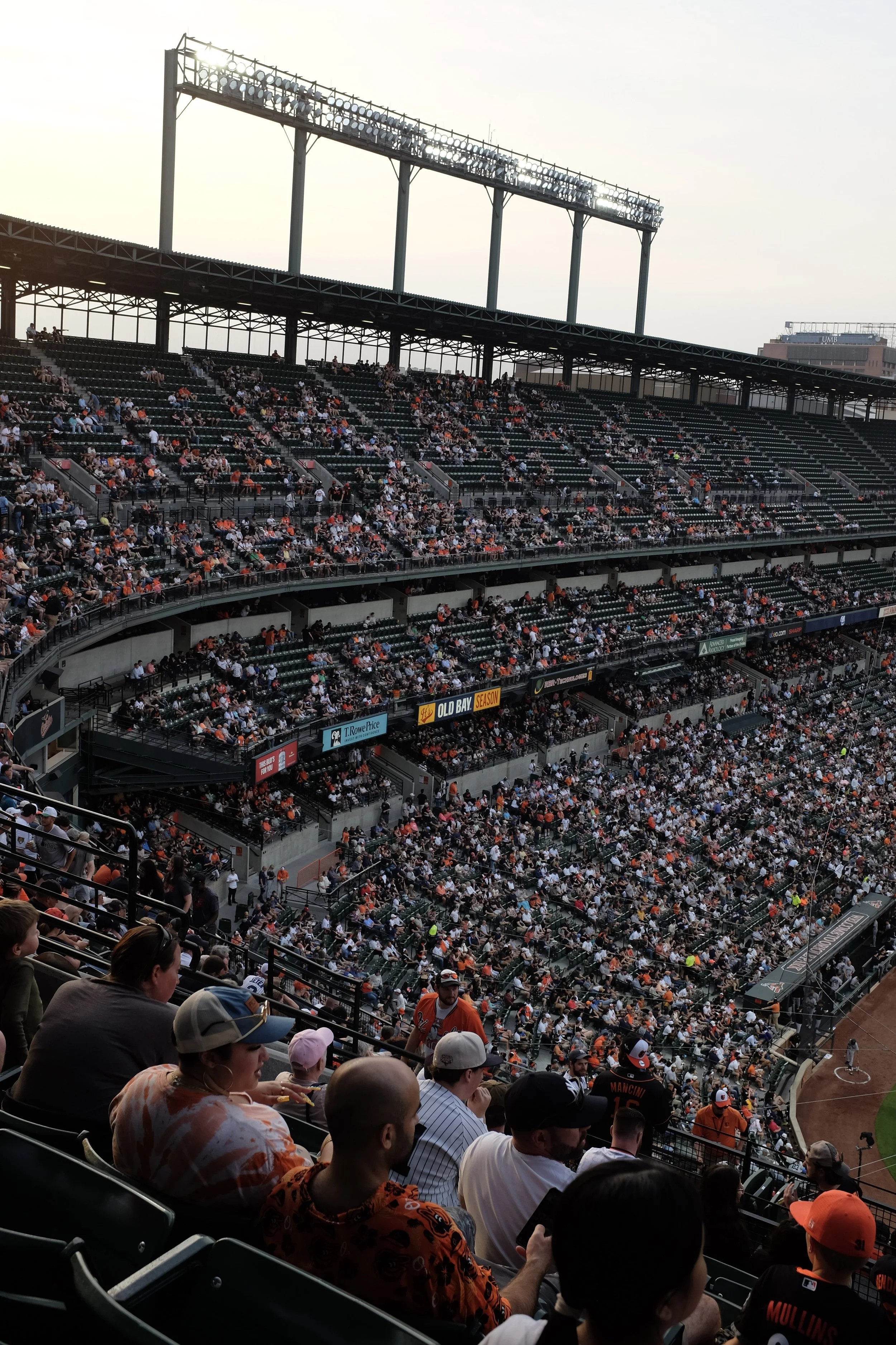 A crowded baseball stadium with spectators seated in the stands, some wearing team jerseys and hats, during daytime.