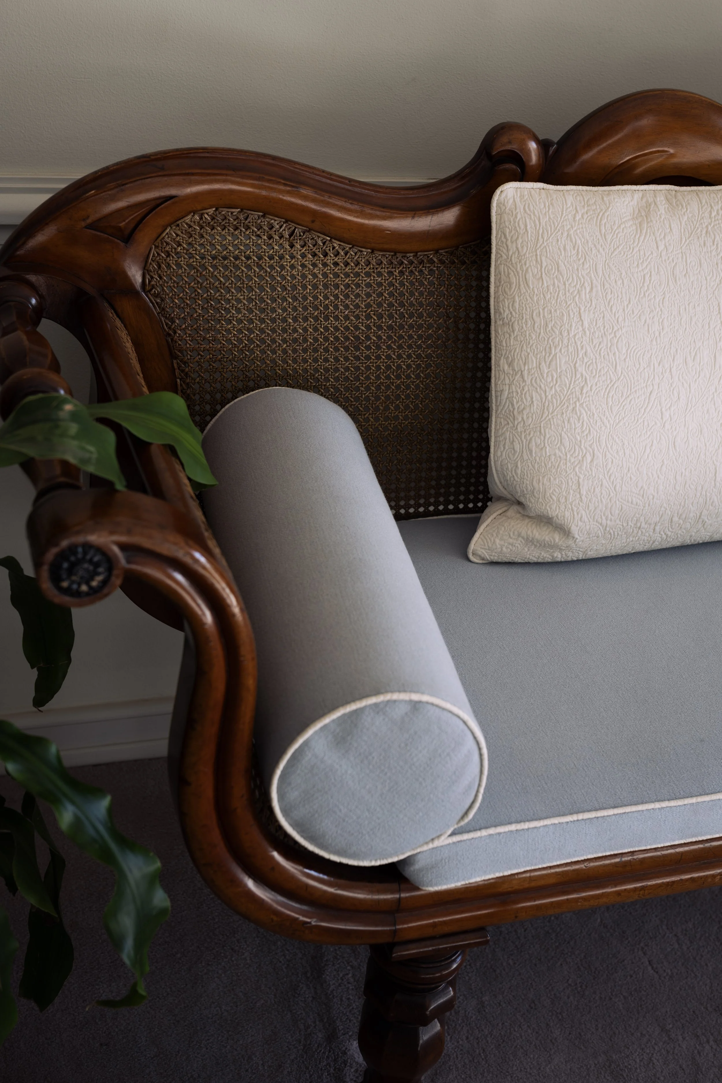 Close-up of a vintage wooden sofa with a woven cane backrest, grey upholstery, a rolled armrest, and a cream decorative pillow.
