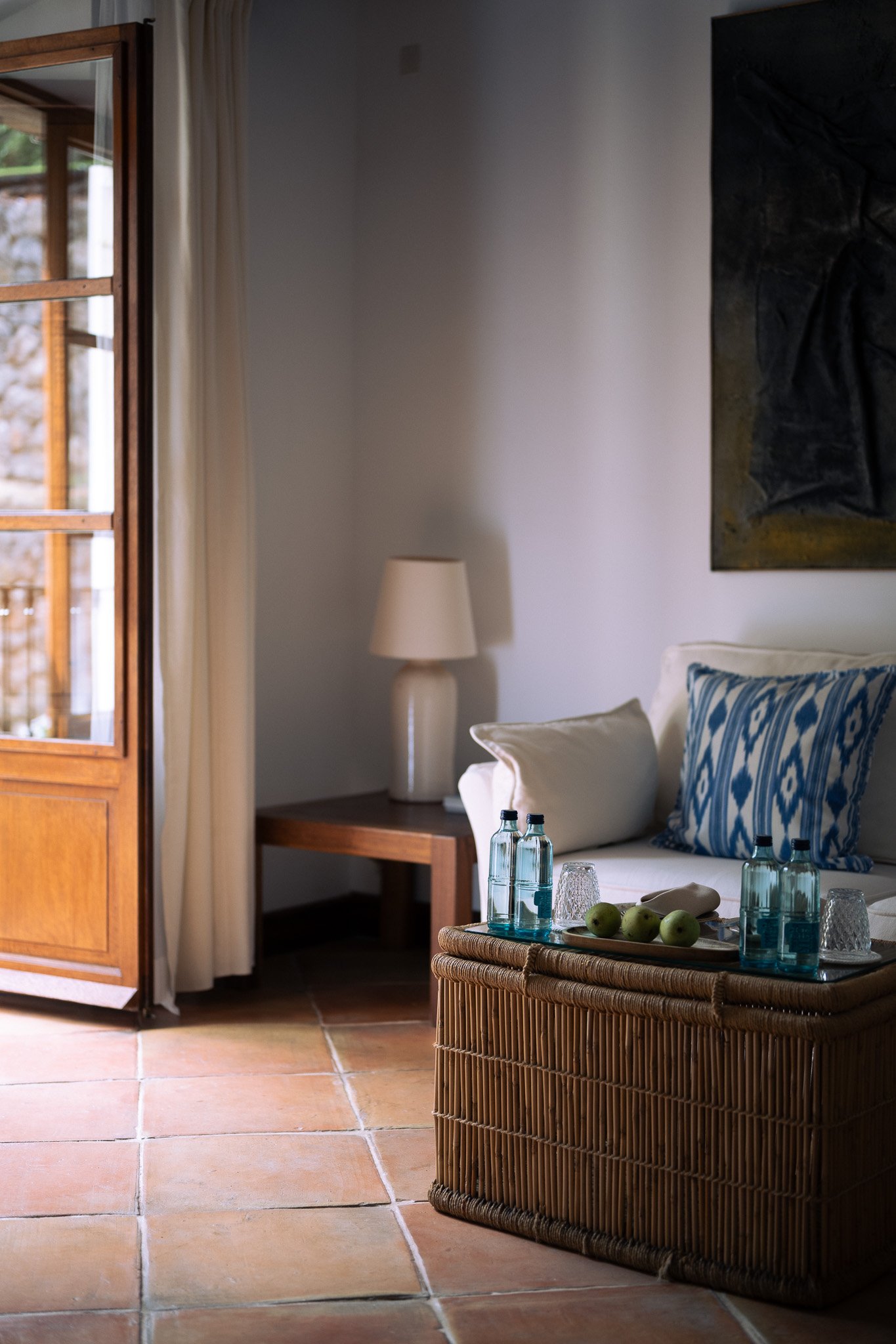 Sunlit guest room with wicker table, blue glass bottles, pears and an open balcony door at La Residencia.