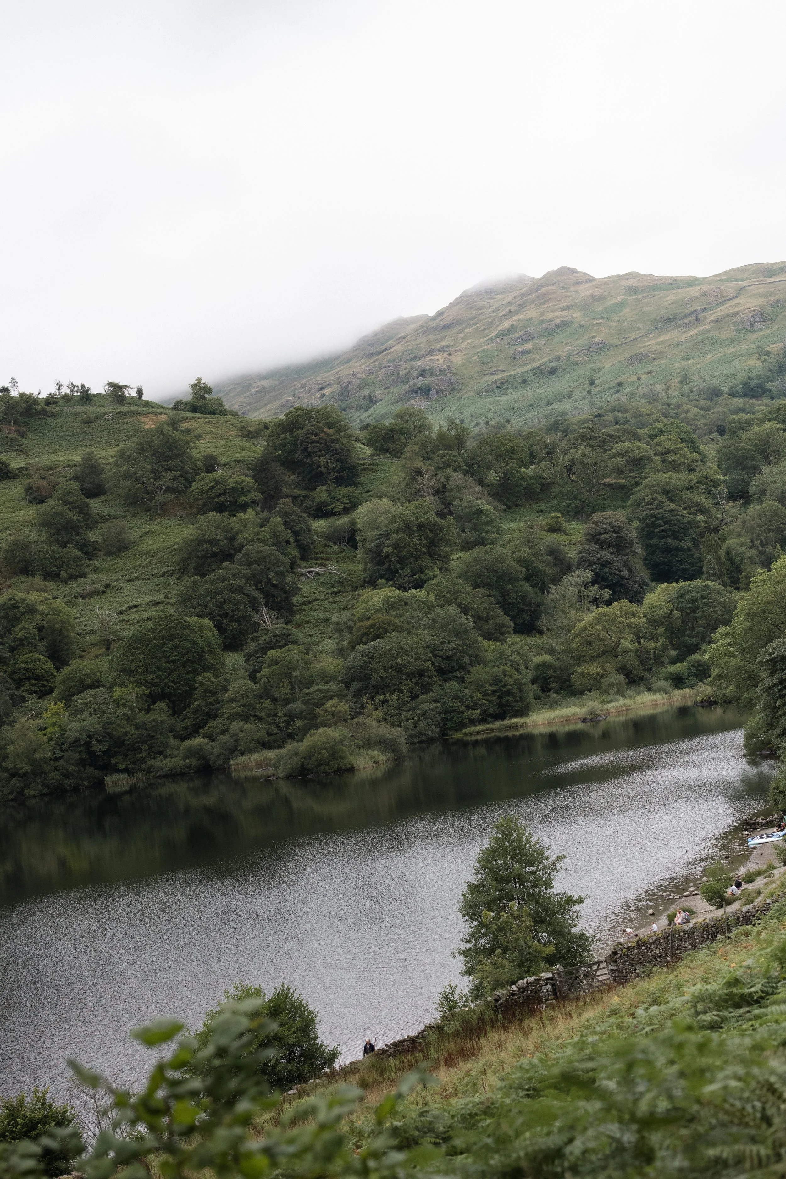 Mist-covered Lake District fells rising above a quiet lake and wooded landscape near Windermere, Cumbria.