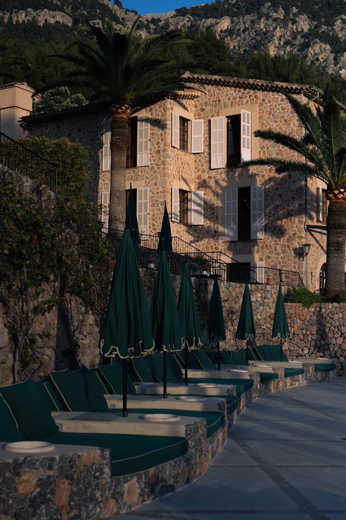 Stone façade with white shutters, palm trees and poolside loungers at La Residencia in Deià, Mallorca.