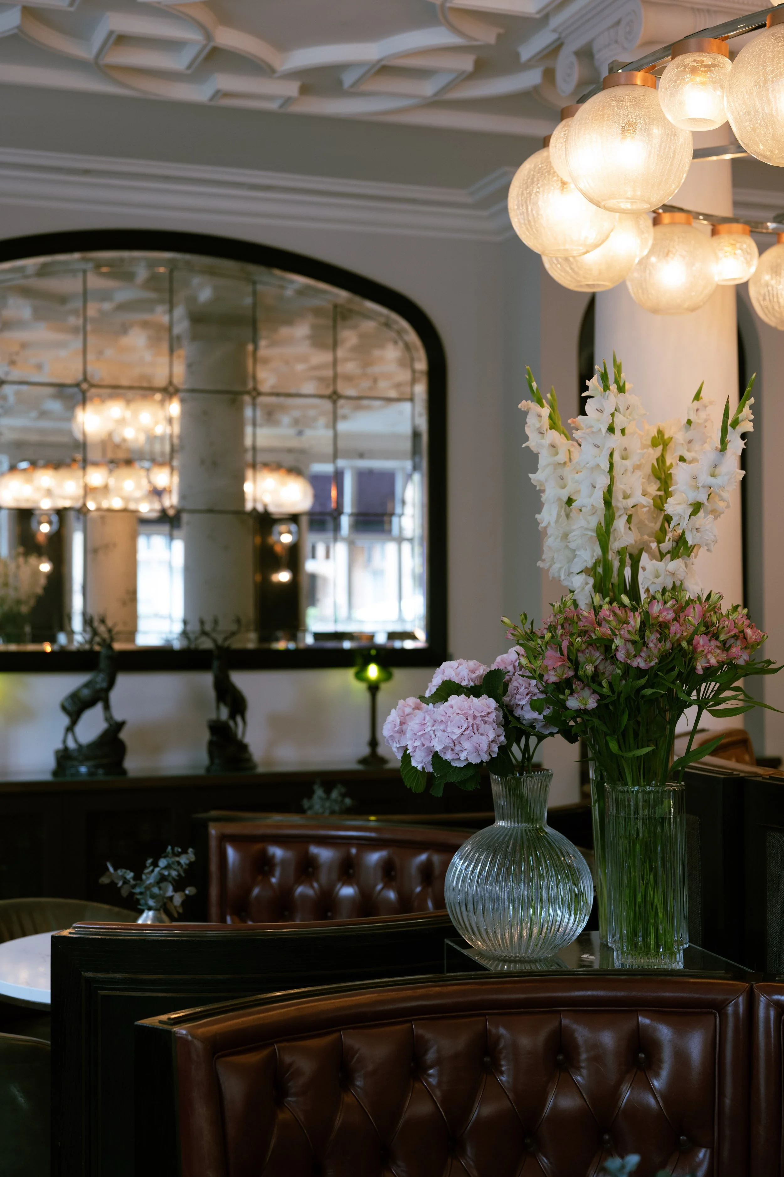 Elegant interior of a restaurant or hotel lobby with flower arrangements in glass vases, vintage leather chairs, a large mirror, and soft lighting fixtures.
