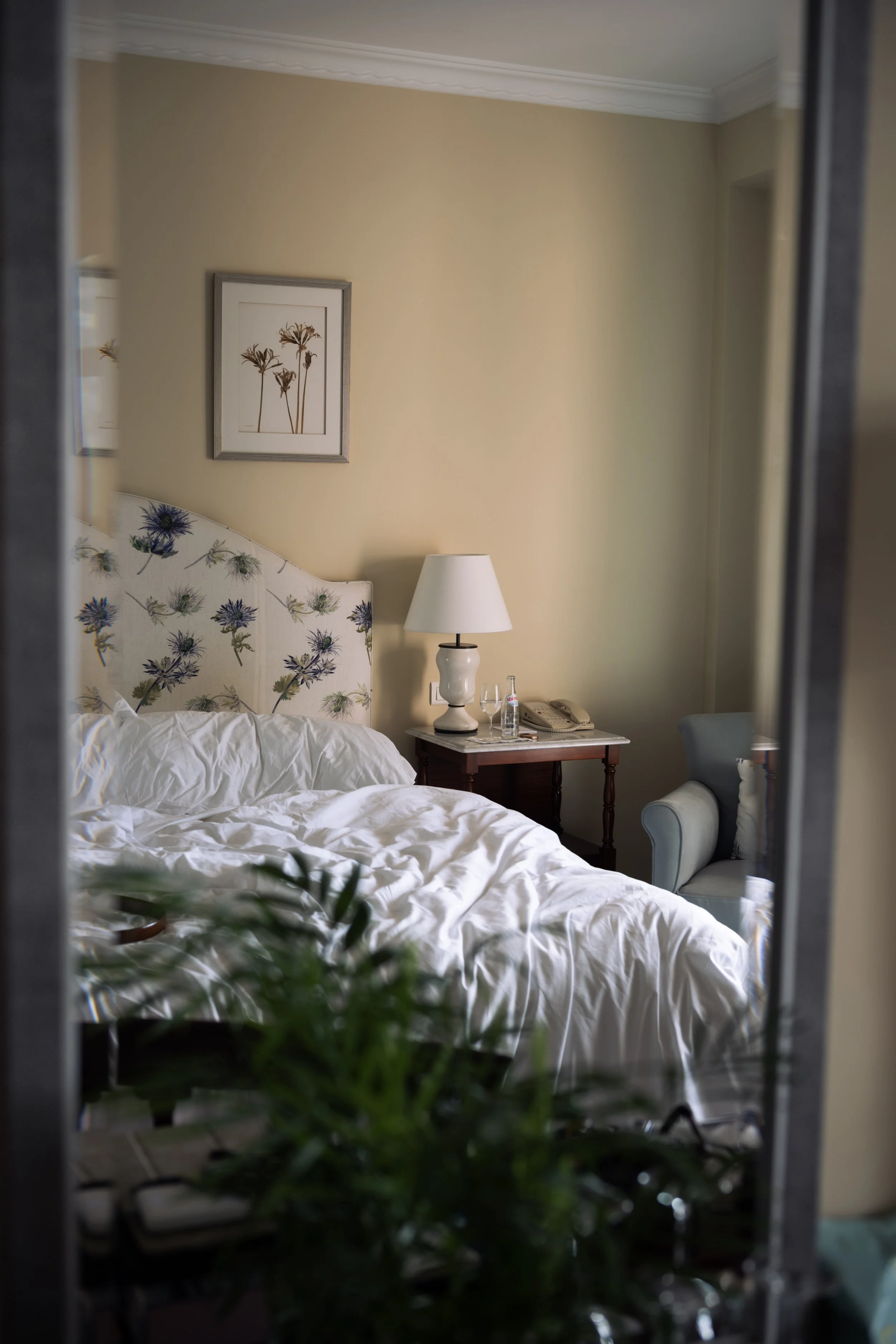 Reflection of a bedroom in a mirror showing a bed with a floral headboard, white bedding, a side table with a white lamp, a telephone, glasses, and a water bottle, with a framed picture of flowers on the wall and a blue armchair.