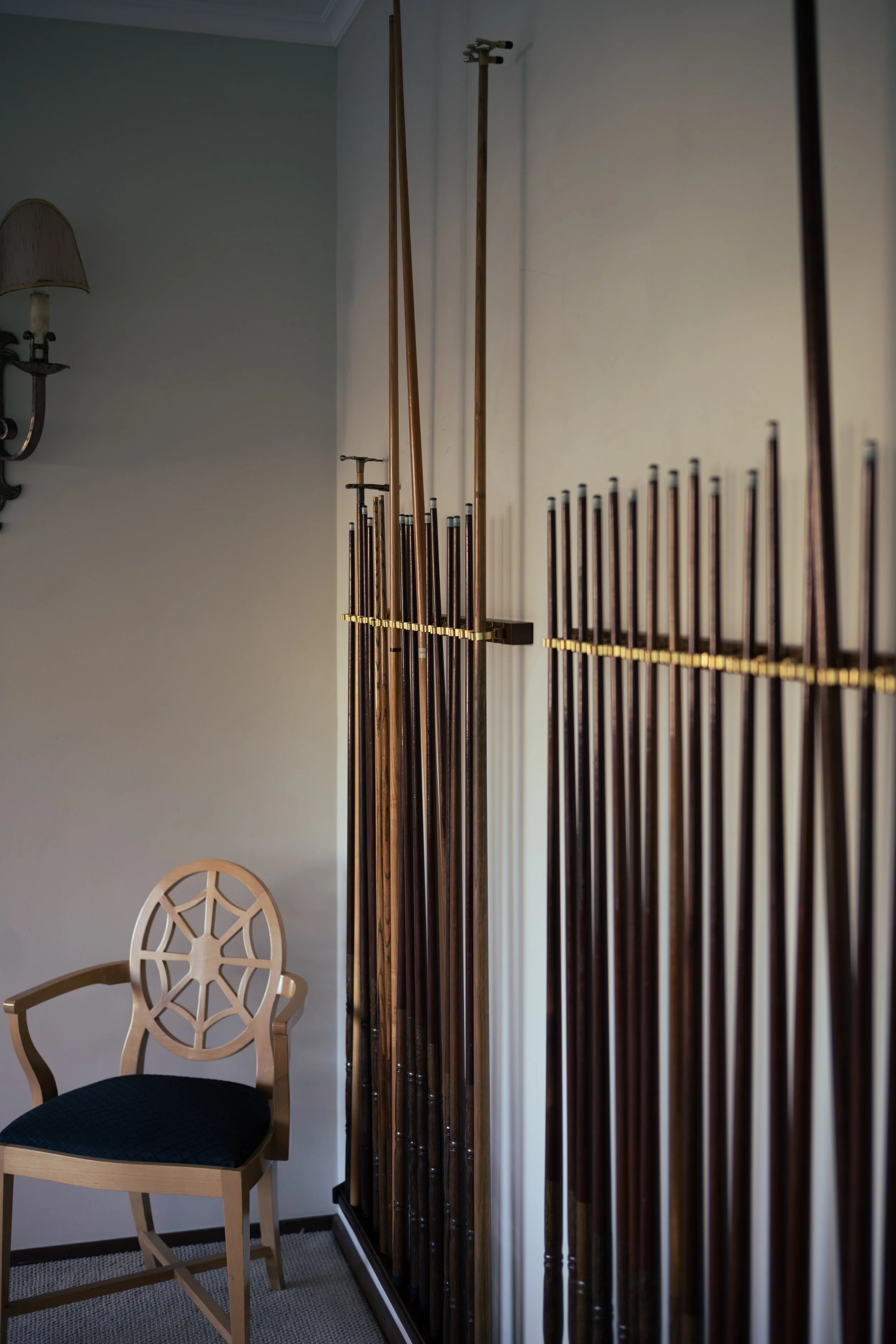 A corner of a room with a wooden chair with a decorative backrest and black cushion, and a collection of wooden cue sticks stored vertically on a rack against the wall.