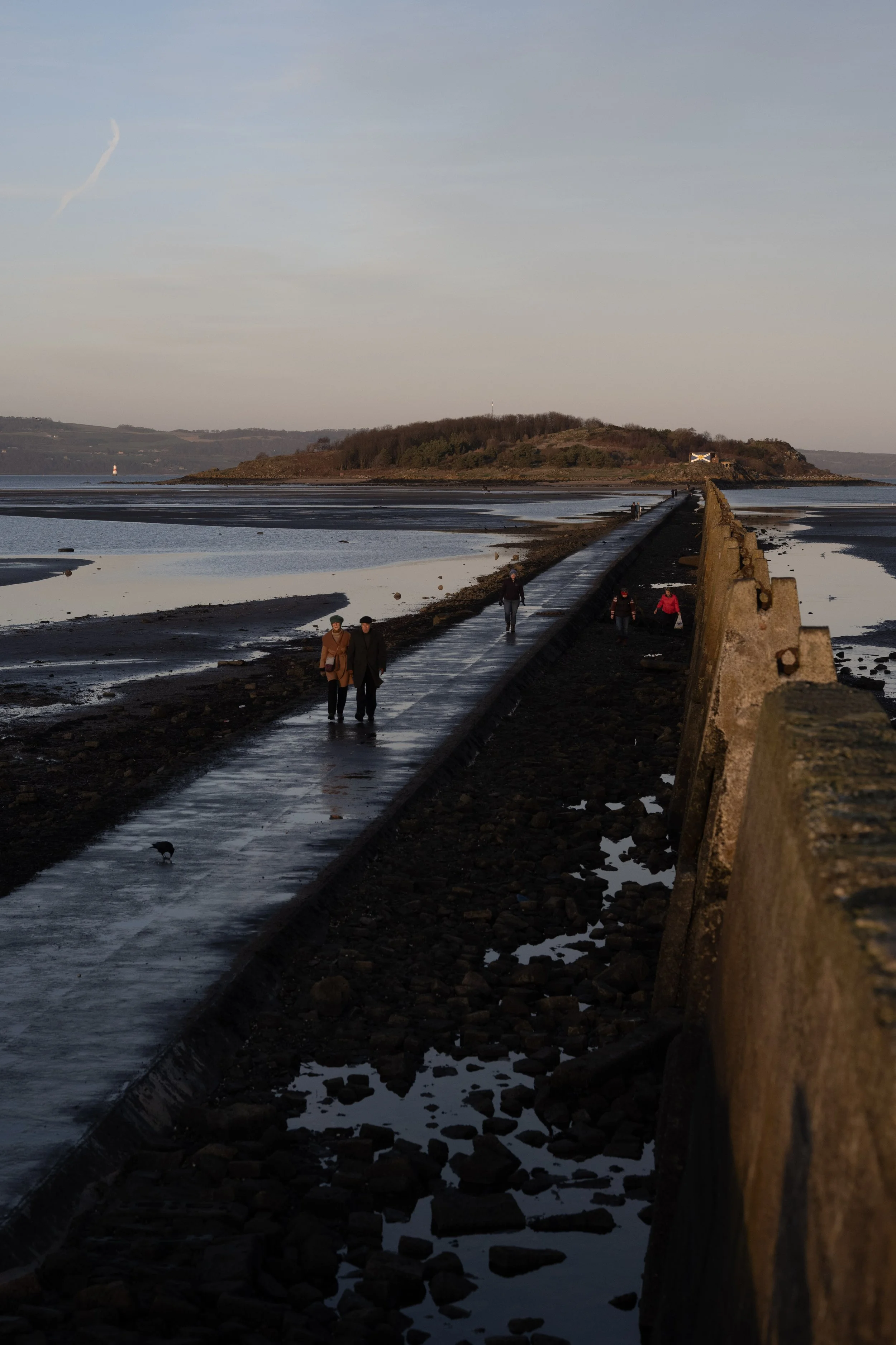 People walking along a narrow pier extending across a tidal flat towards a small island with trees and a flag, during sunset or early evening.