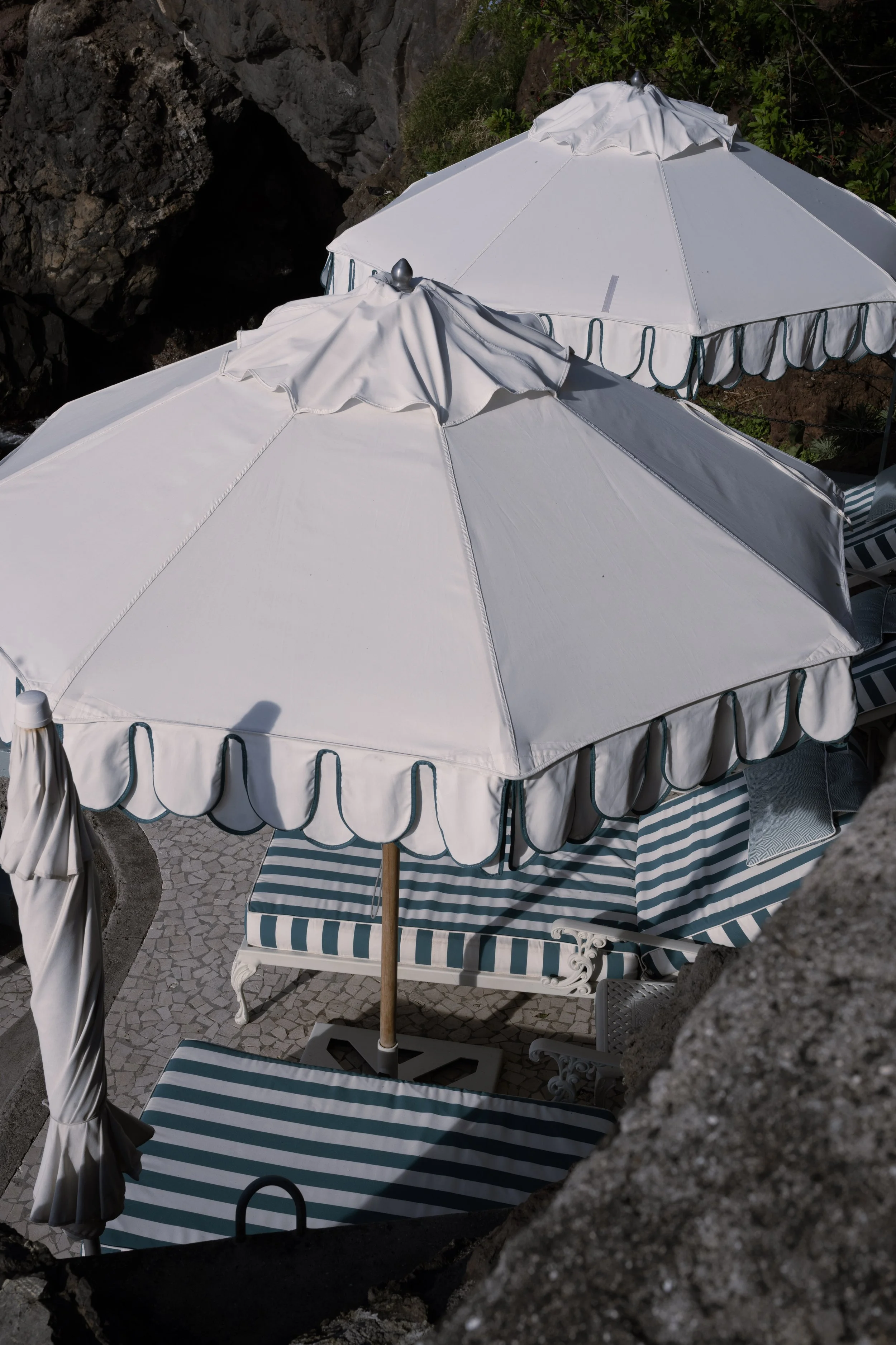 Two white beach umbrellas with ruffled edges shade striped lounge chairs on a stone patio by the rocks and greenery.