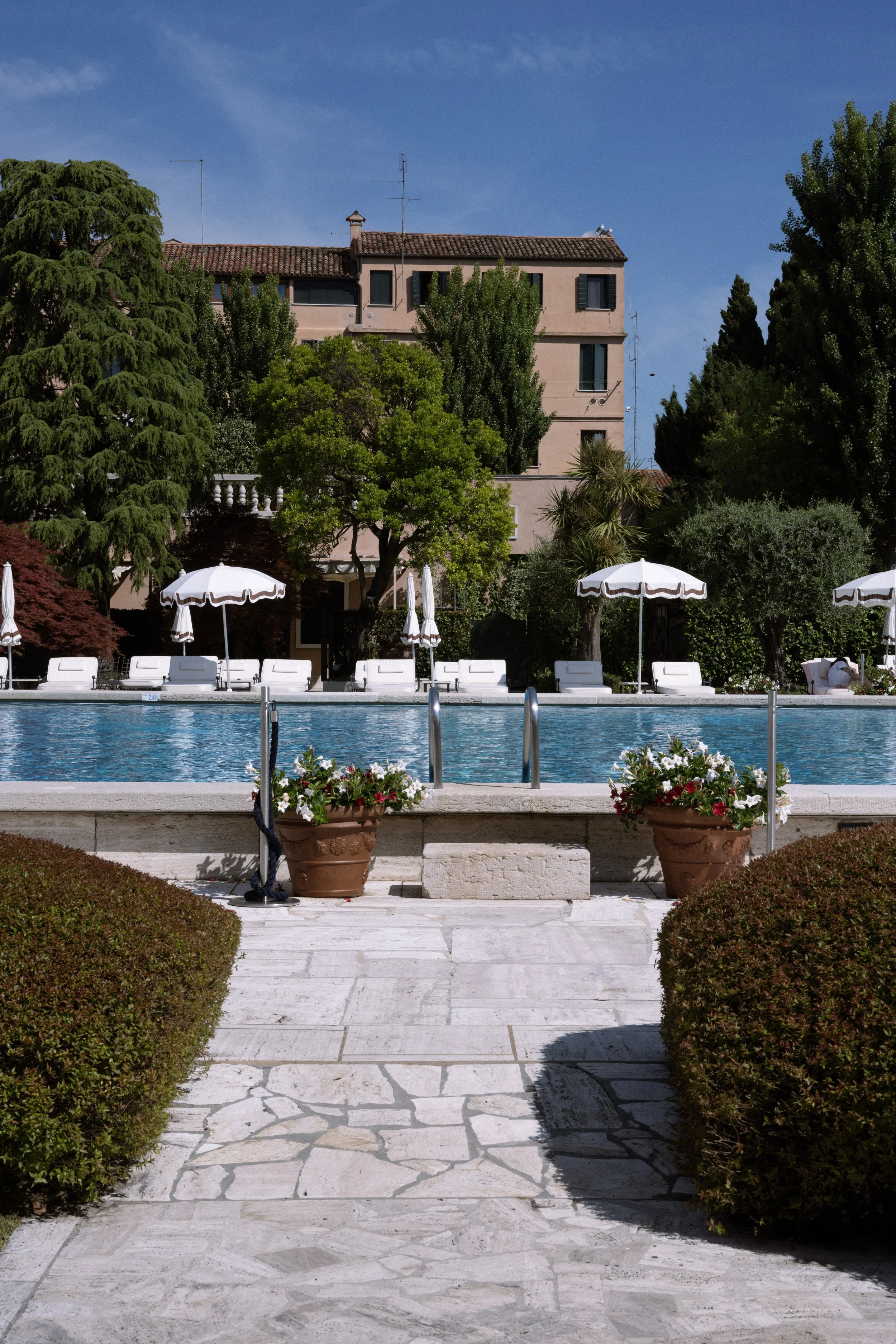 A view of a swimming pool with white lounge chairs and umbrellas, surrounded by greenery, with a pink building in the background under a blue sky.