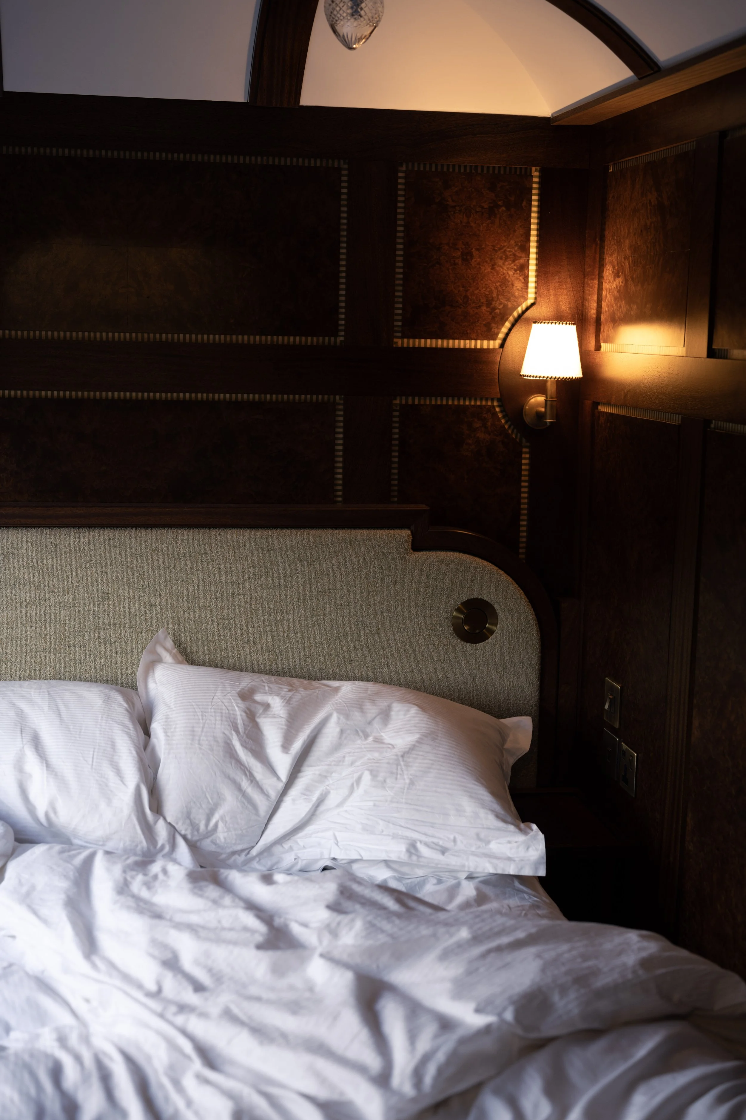 A bed with white sheets and pillows in a hotel room, with a wooden headboard, a wall-mounted lamp, and decorative wood paneling on the wall.