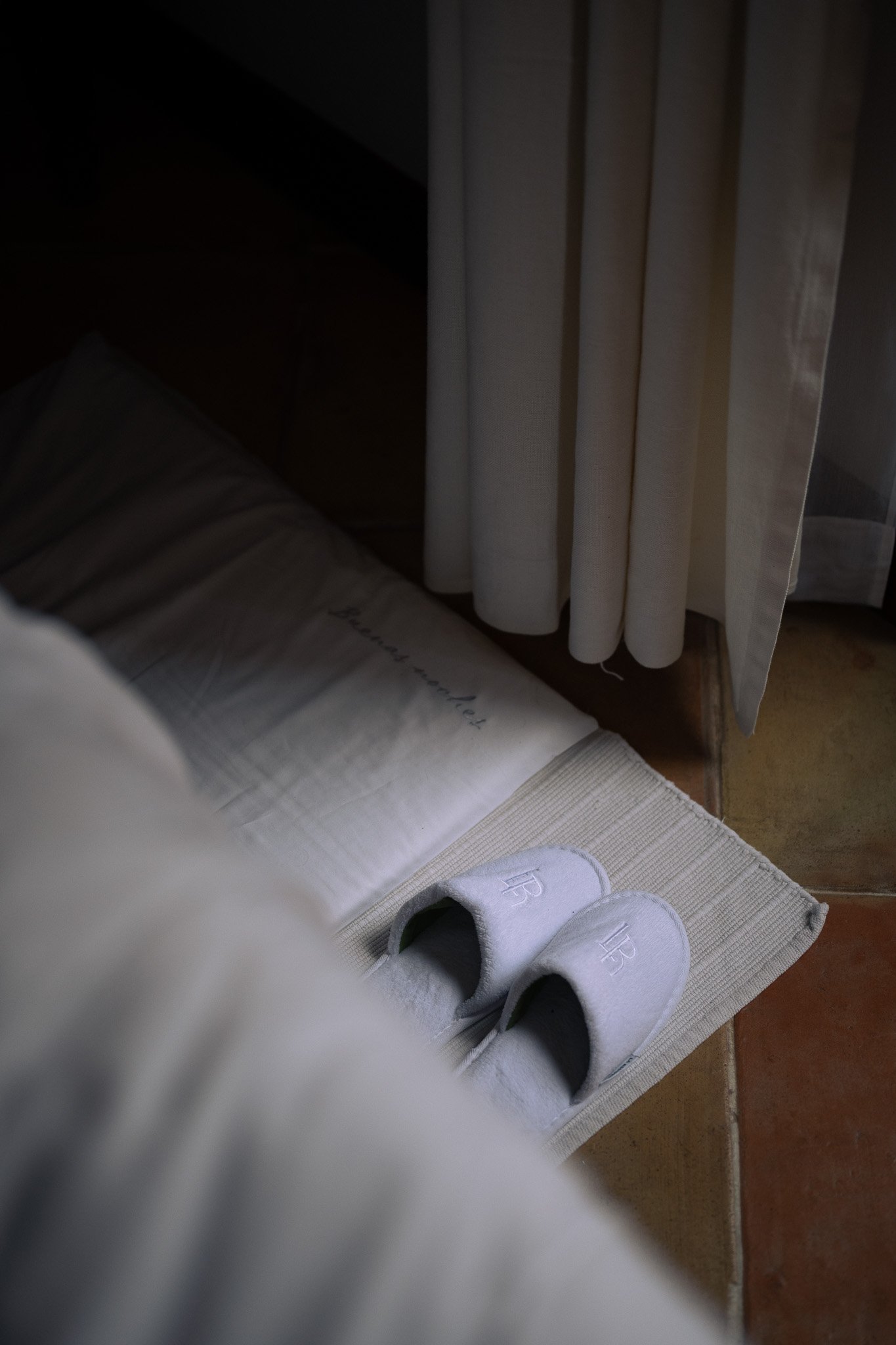 White La Residencia slippers beside a four-poster bed and sheer curtains in a guest room.