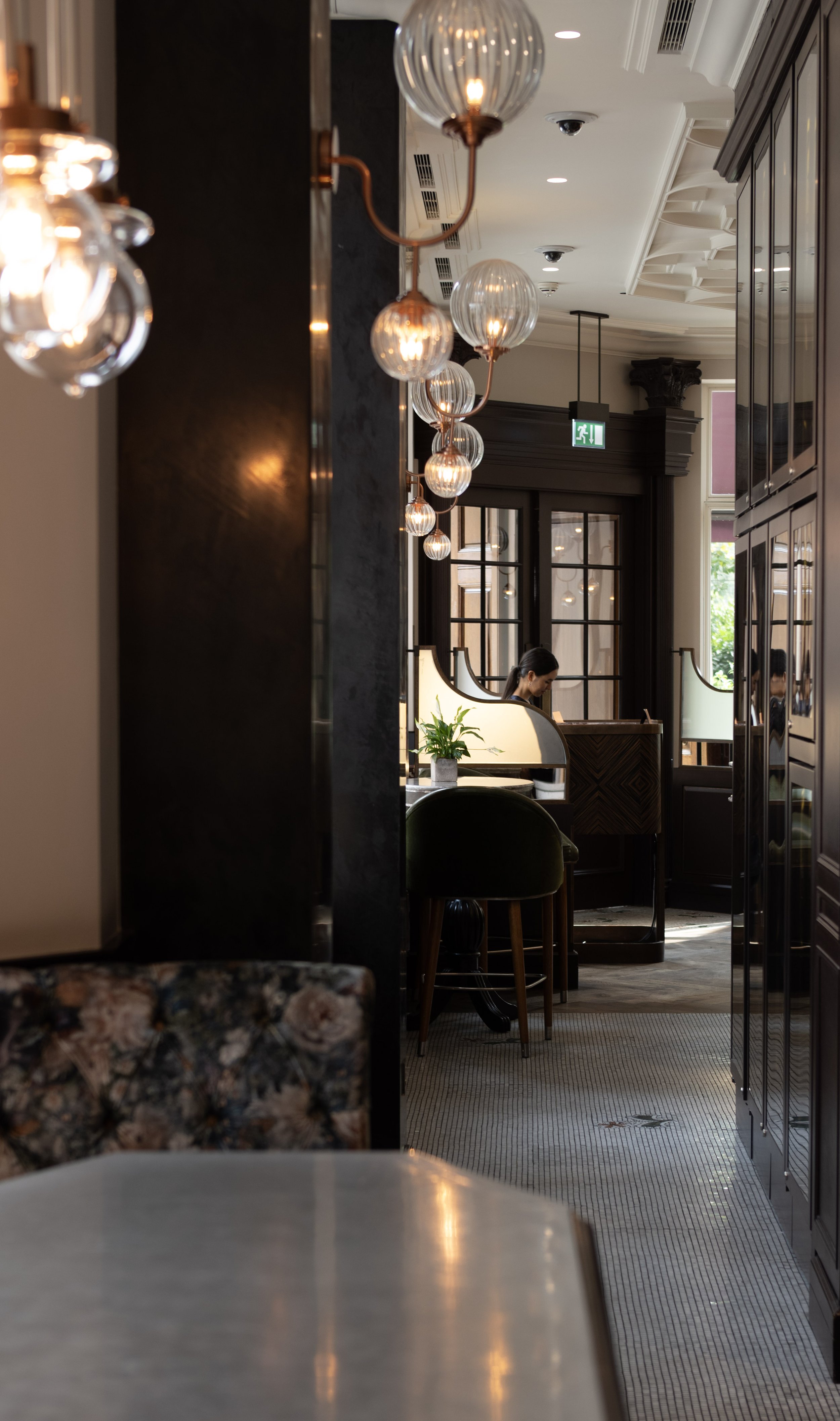 A hotel lobby area with elegant hanging globe lights, dark wooden furniture, and a woman seated at a desk near windows. The scene includes a partial view of a marble table in the foreground and potted plant next to the desk.