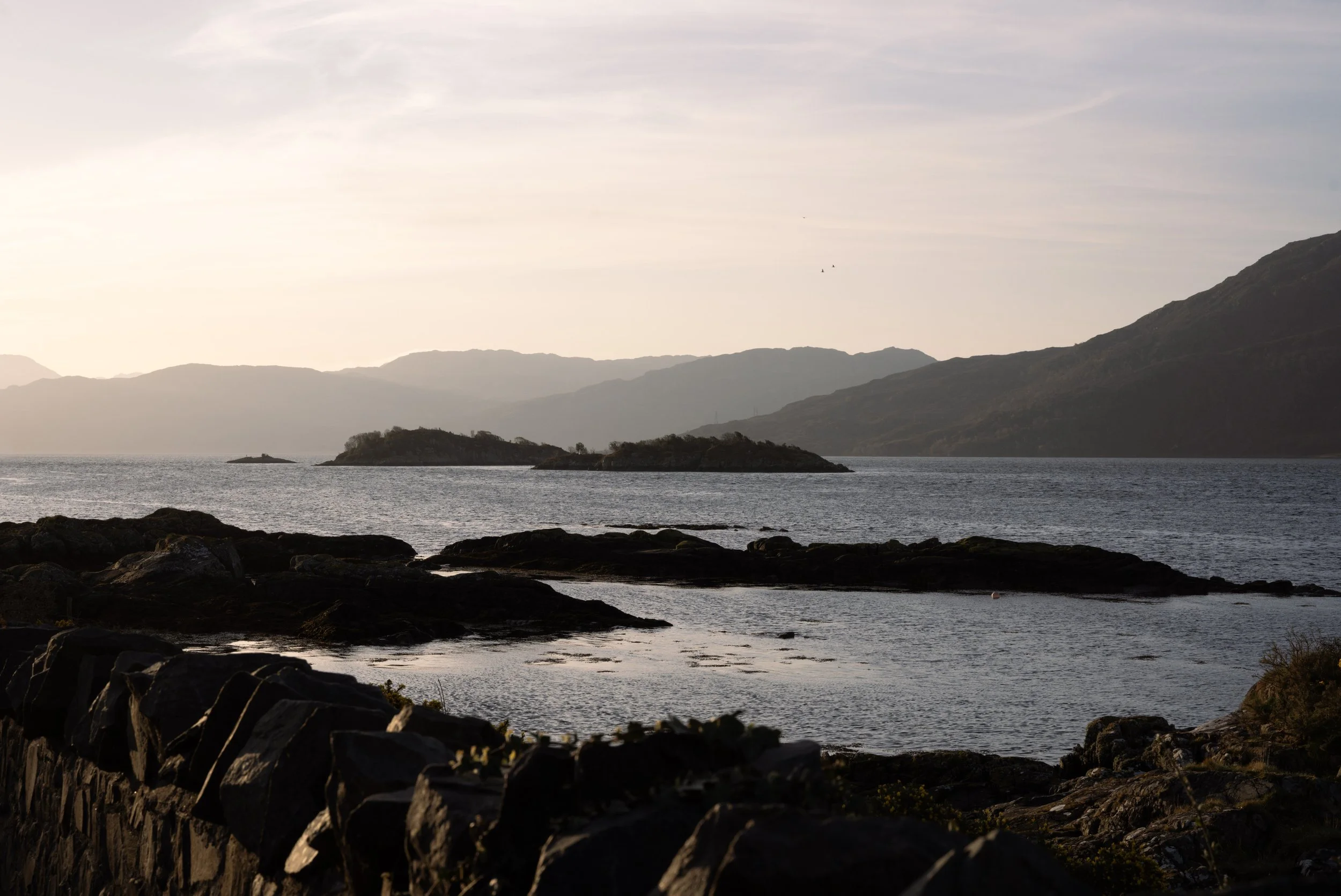 Scenic view of a lake with small islands, rocky shoreline in the foreground, and mountains in the background during sunset.