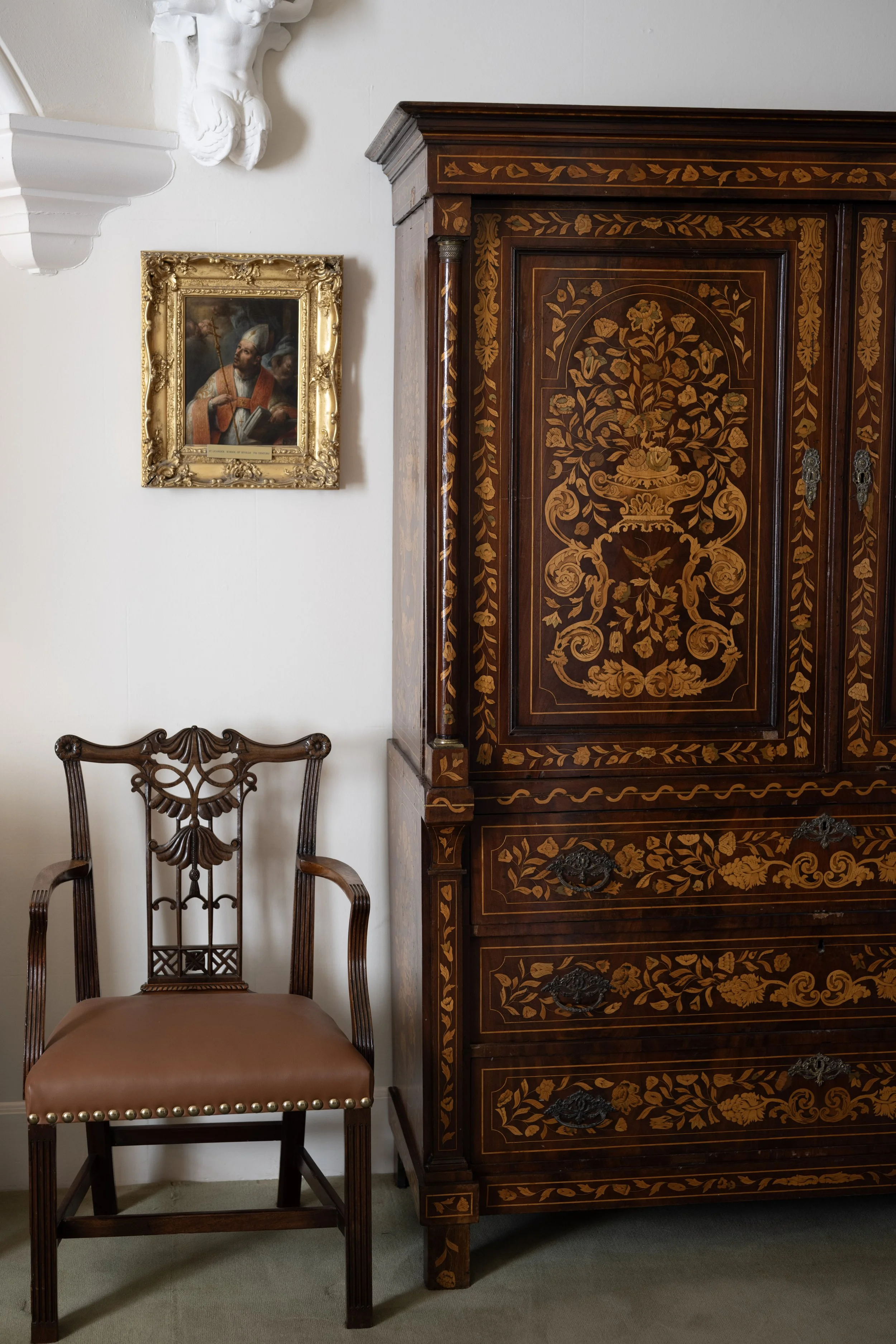 A wooden antique chair with a carved backrest and leather seat, a tall wooden cabinet with intricate floral inlay, a framed portrait of a man in historical military attire on the wall, and decorative white sculptures on the wall.