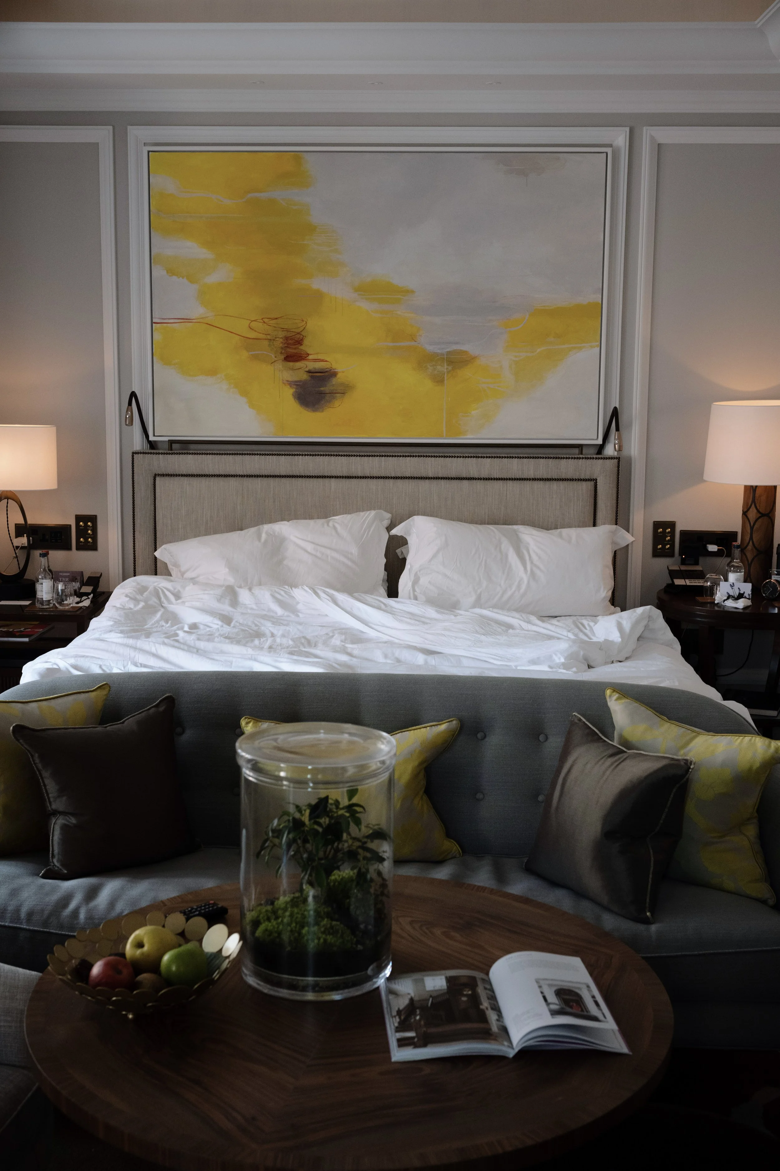 A hotel room with a bed against the wall, covered with white bedding, and pillows. There are two nightstands with lamps on each side of the bed. A wooden coffee table in front has a glass terrarium with plants, a bowl of fruit, and an open magazine. 