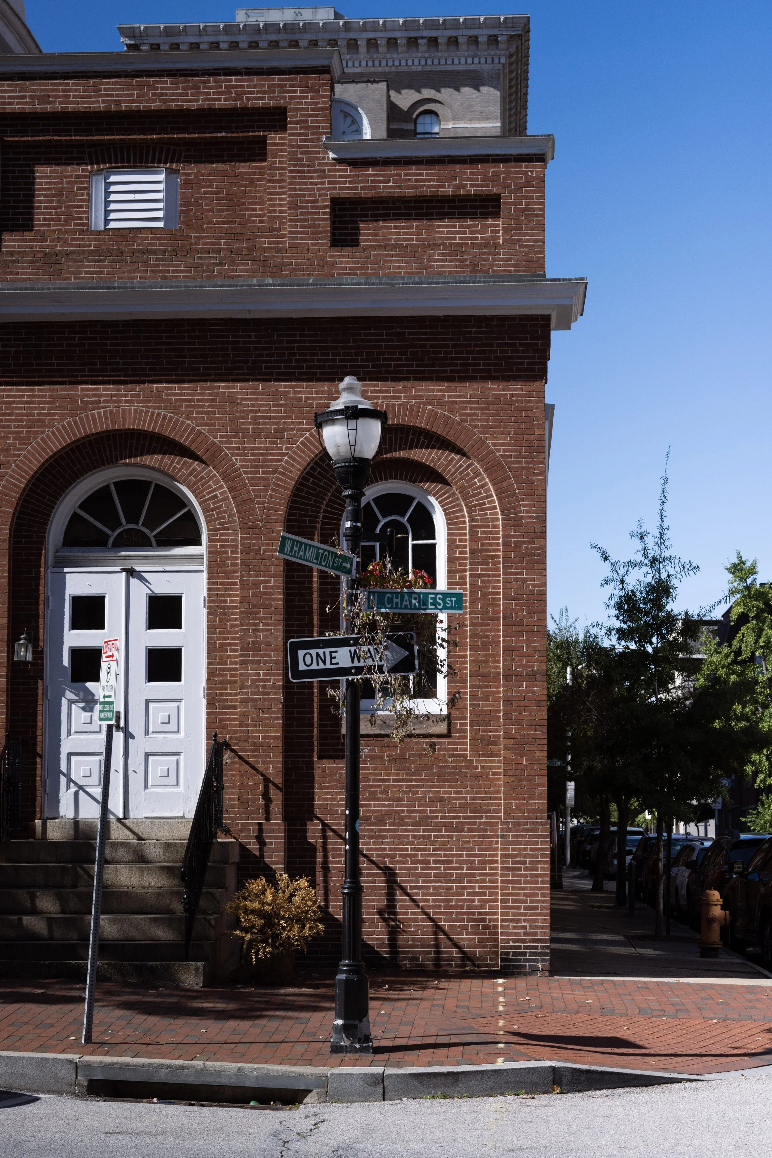 Street corner with a lampposts, street signs, a brick building, stairs leading to a white door, and a tree on the right side under a clear blue sky.