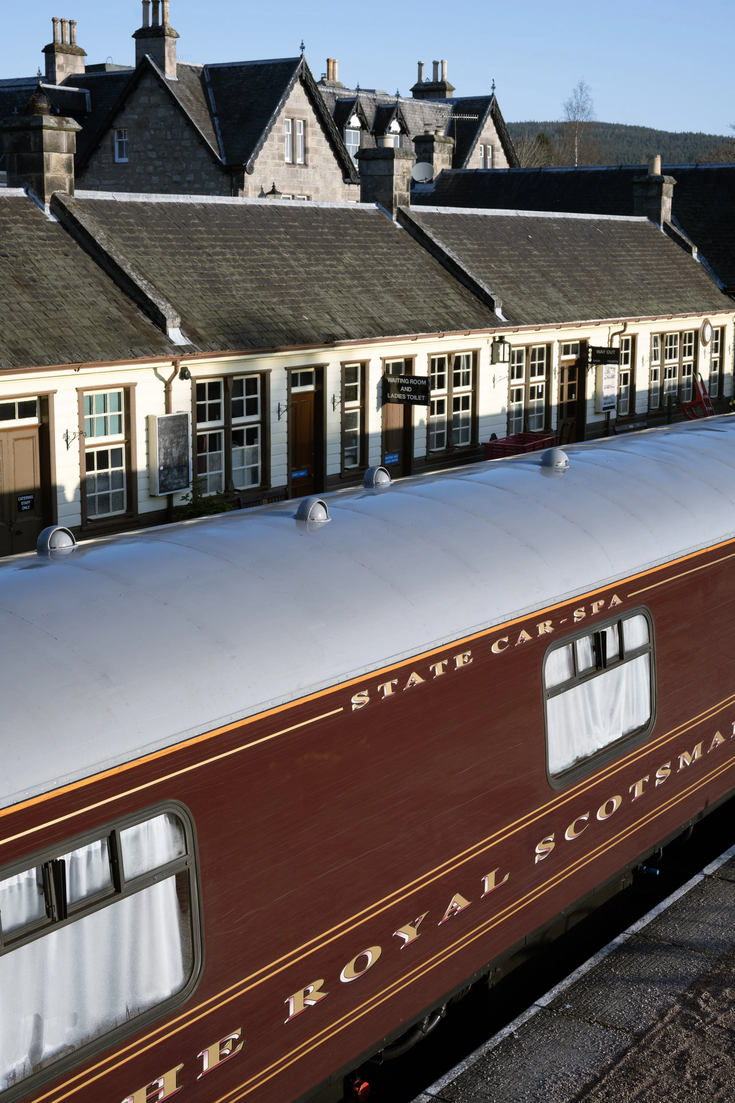 A vintage train car with the text 'The Royal Scottish_Mail' is parked on a station platform. Behind the train, there are buildings with pitched roofs and chimneys, and signs indicating waiting rooms and ladies' toilets.