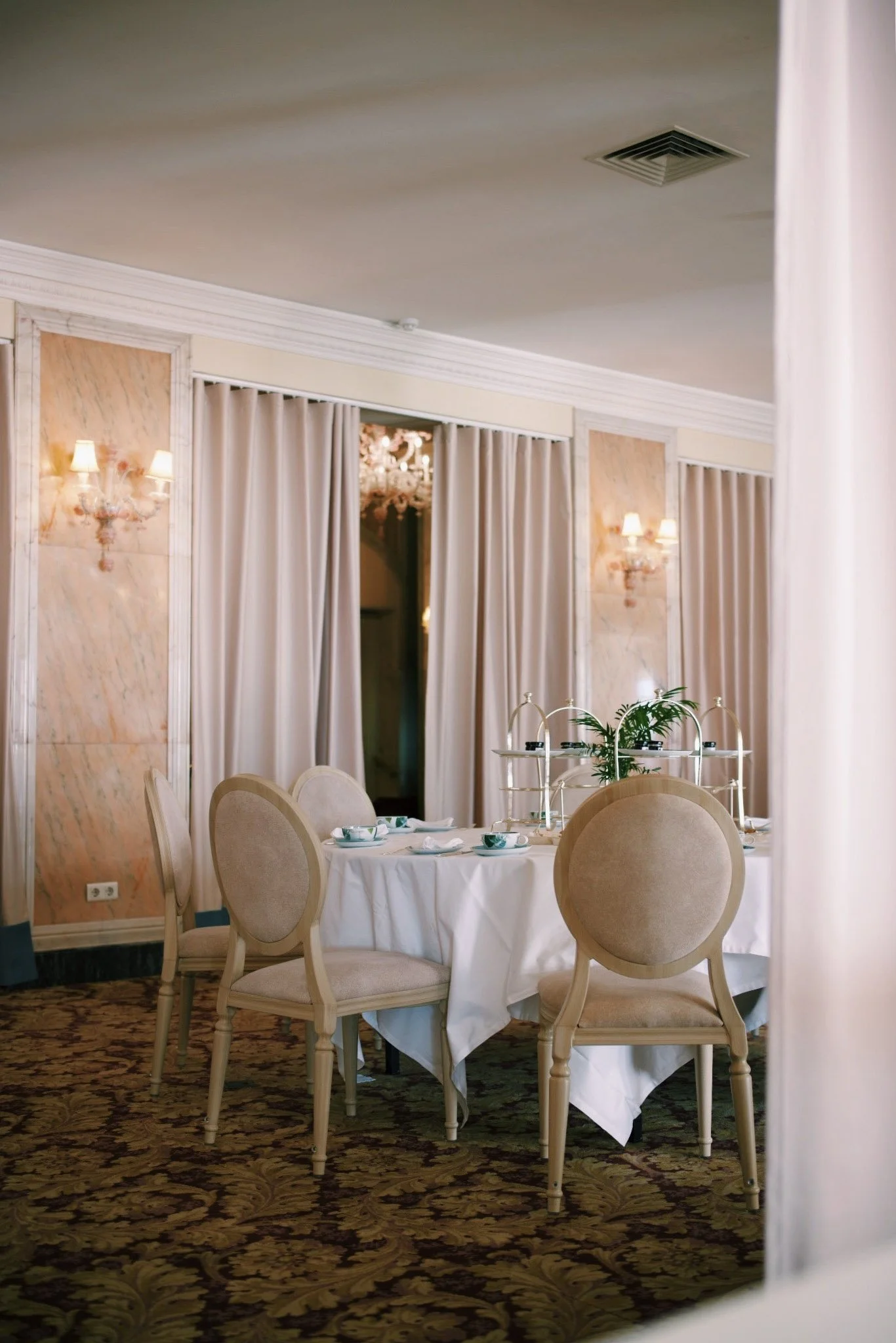 An elegantly set dining table with six beige upholstered chairs in a decorated room with beige curtains, wall lamps, and a chandelier visible through the curtains.