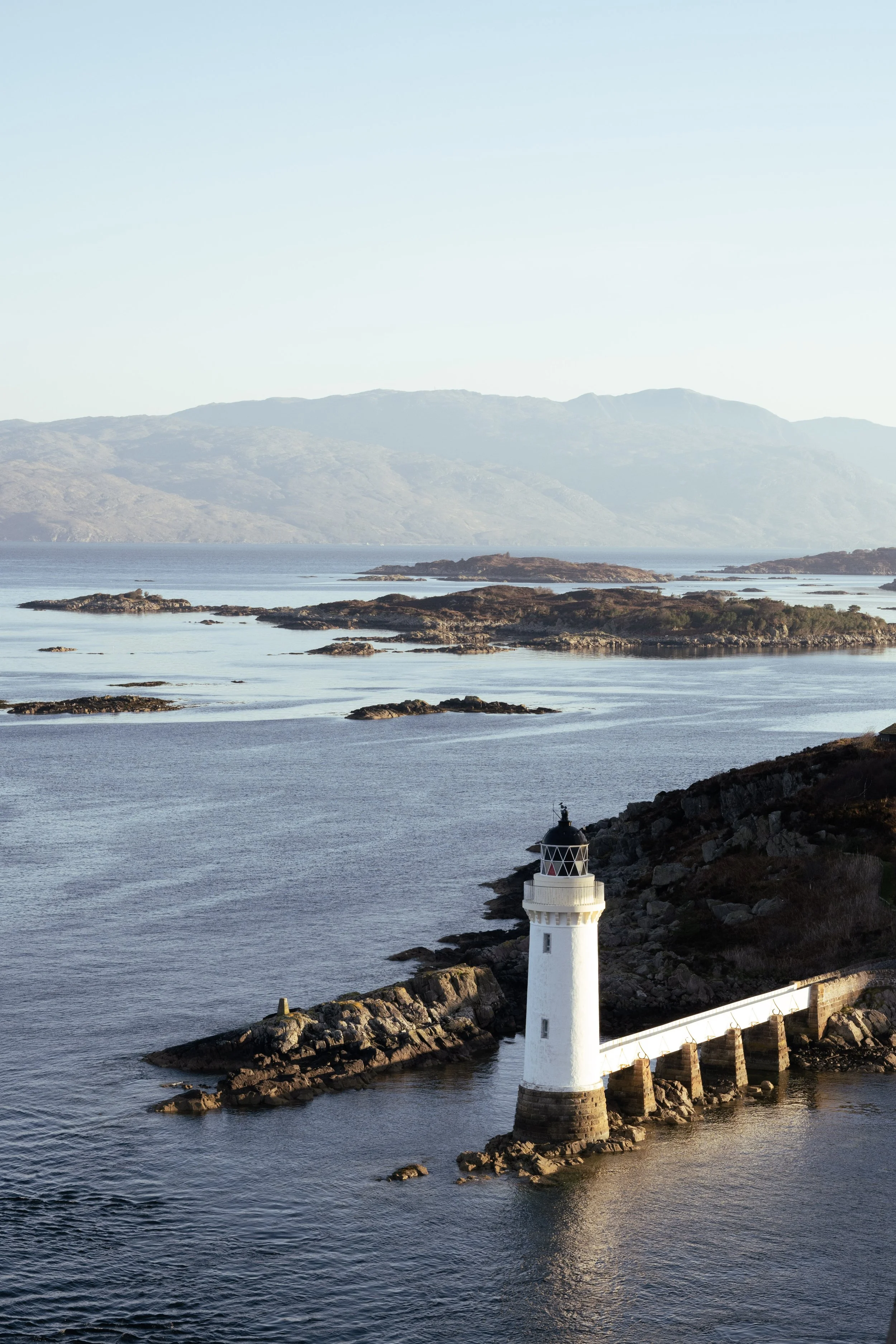 Lighthouse on rocky shoreline with calm sea, small islands, and distant mountains under clear sky.