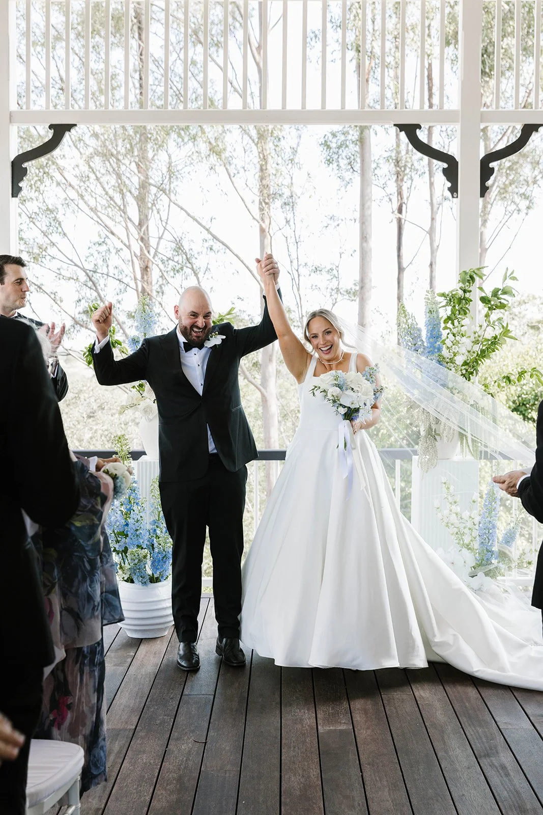 Outdoor wedding ceremony by the ocean with a bride and groom under a floral arch, surrounded by bridesmaids, groomsmen, and seated guests.