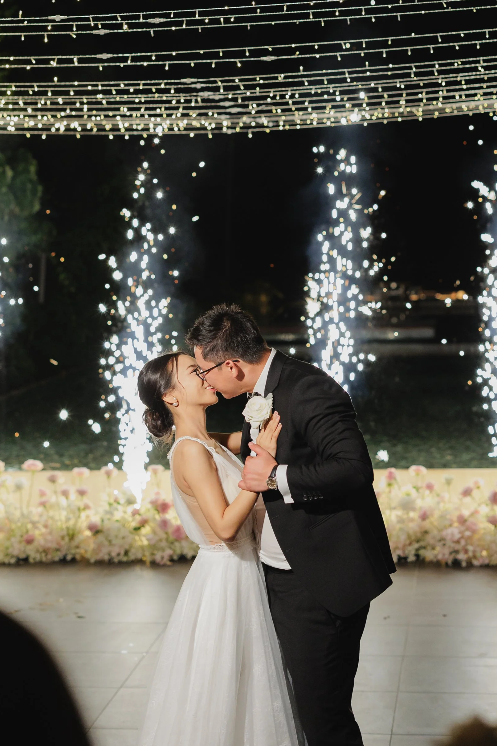Bride and groom celebrating outdoors, holding hands, with guests throwing confetti, surrounded by flowers and trees.