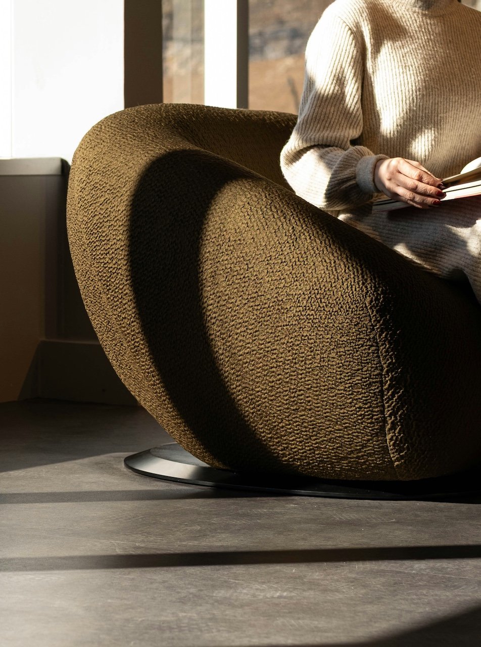 Close-up of a person sitting in a textured brown modern armchair, reading a book, with sunlight casting shadows in a well-lit room.