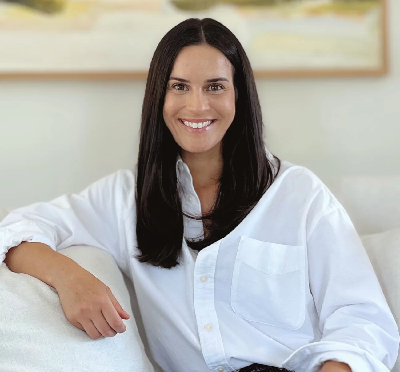 A woman with long dark hair and a white shirt smiling and sitting on a white couch with a blurred painting in the background.