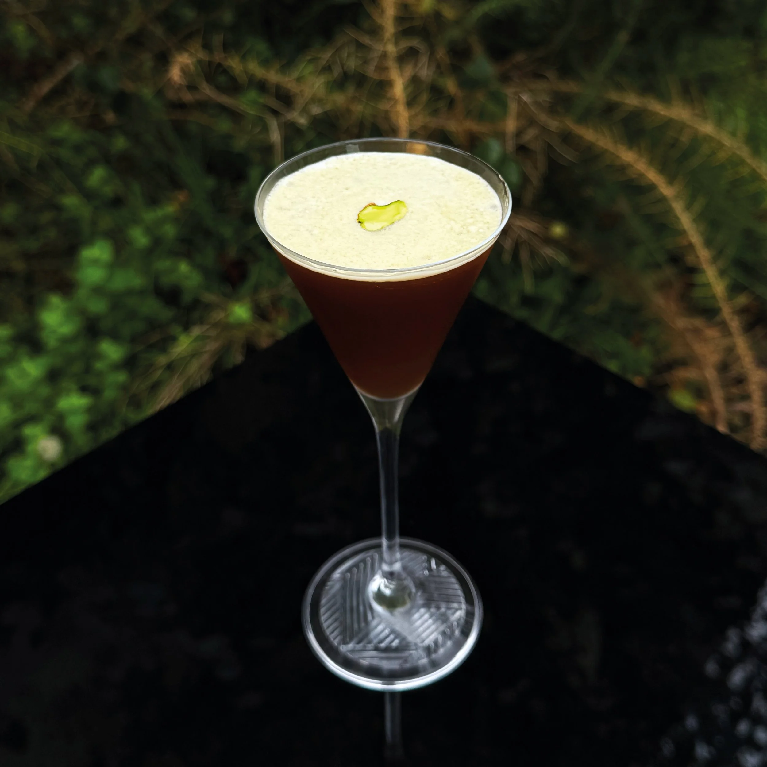 A STRATO cocktail in a martini glass topped with green garnish and placed against a blurred outdoor background.