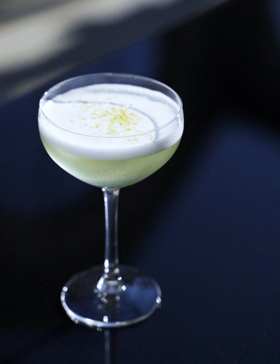 A cocktail glass with a pale green STRATO drink topped with a layer of white foam and a sprinkle of lemon zest, set against a dark background.
