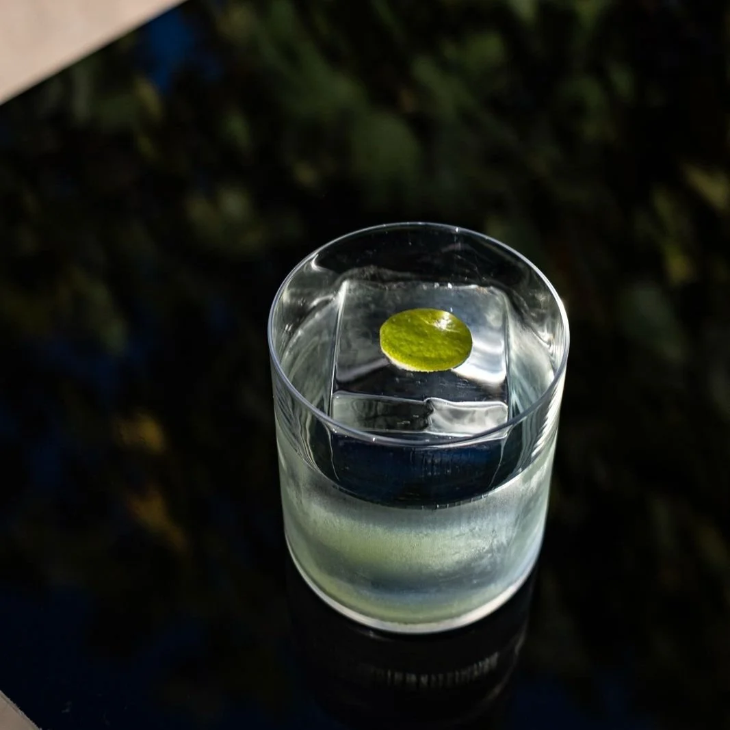 A glass of clear STRATO cocktail with an ice cube and a small green lime wedge inside, set on a dark surface with a blurred background.