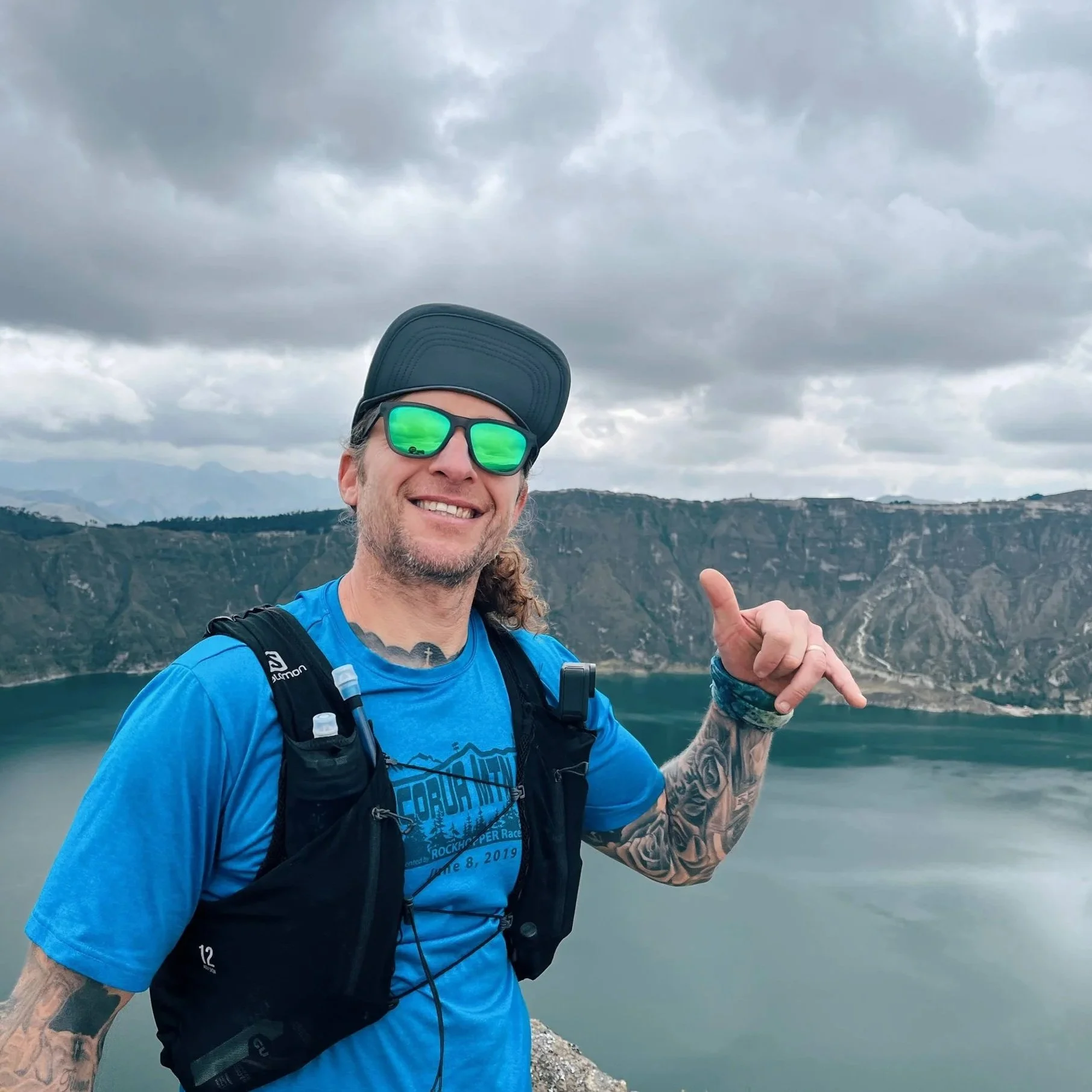 Man wearing sunglasses and a blue T-shirt, smiling and pointing, standing in front of a volcanic crater lake with cloudy sky.