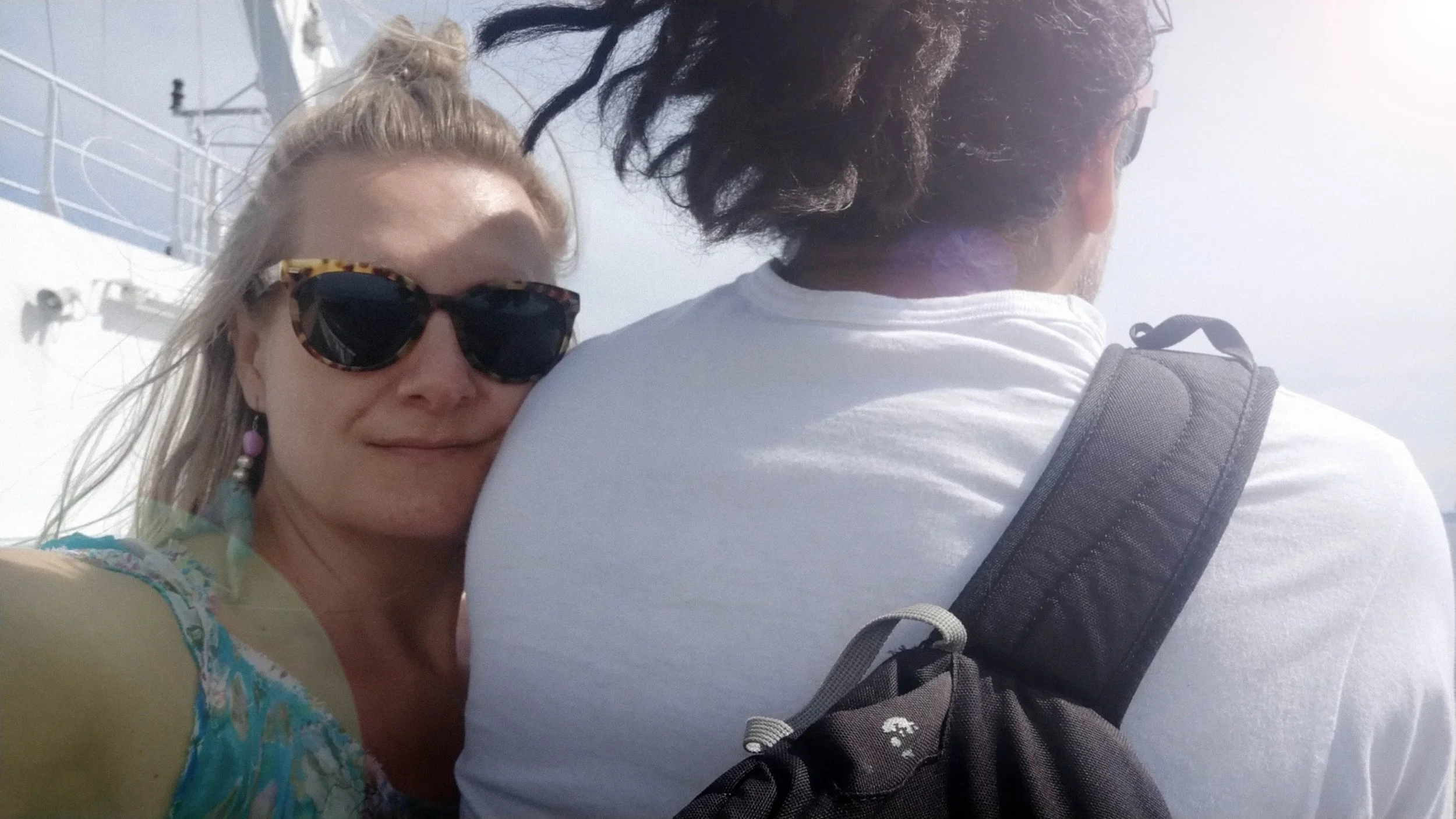 A woman with blonde hair wearing sunglasses and earrings, resting her head on a man's shoulder on a boat.