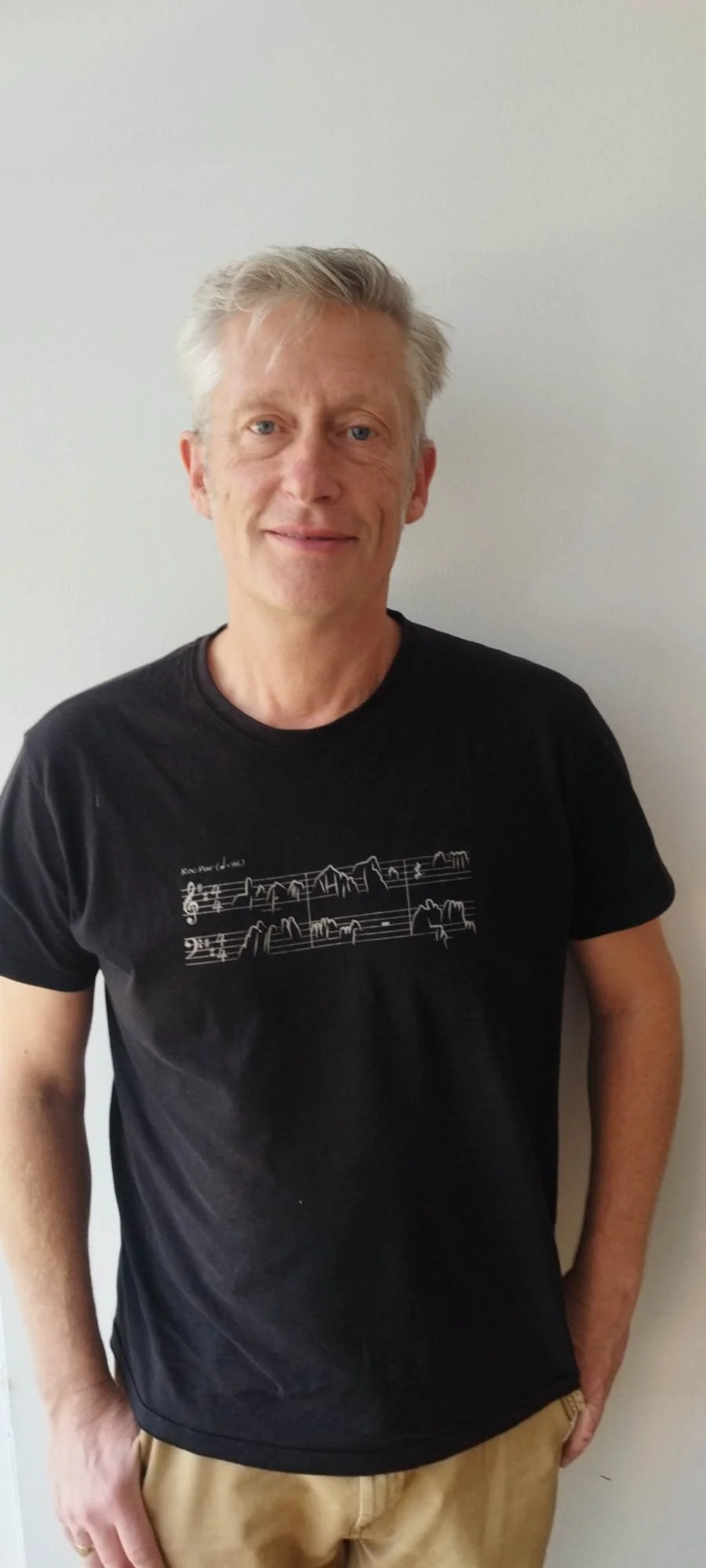 A man with gray hair wearing a beige T-shirt with bicycles printed on it, standing indoors with art frames on the wall behind him.
