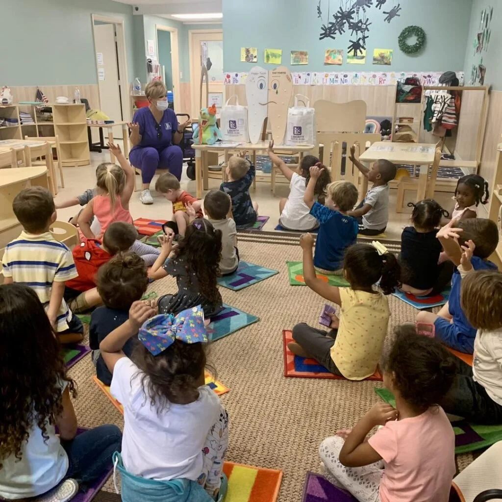 A classroom with a teacher and young children sitting on mats on the floor. The teacher is holding a puppet and wearing a face mask, engaging with the kids during storytime or a lesson. Children are raising their hands and attentively participating.