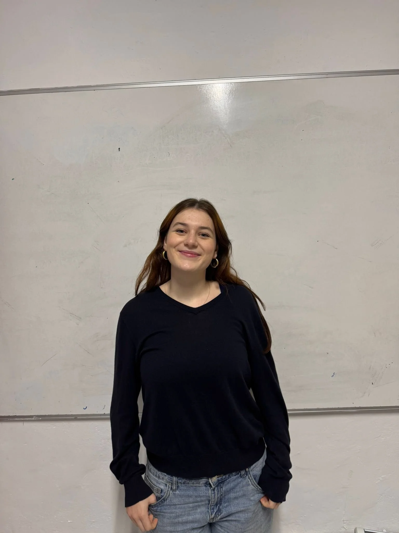 Young man with dark hair and beard standing outside a glass storefront, wearing a white t-shirt.