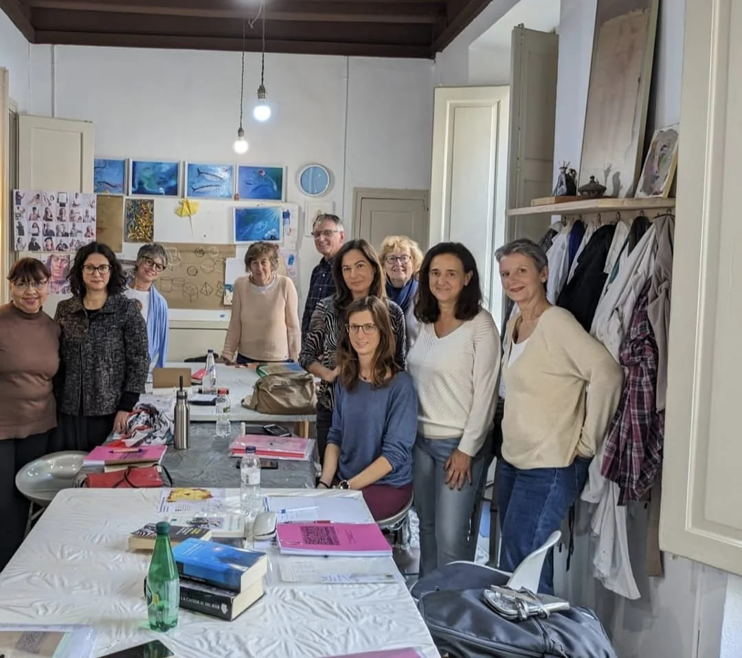 A group of eleven people posing in a room with artwork and supplies, standing and sitting around a table covered with papers, bottles, and books.