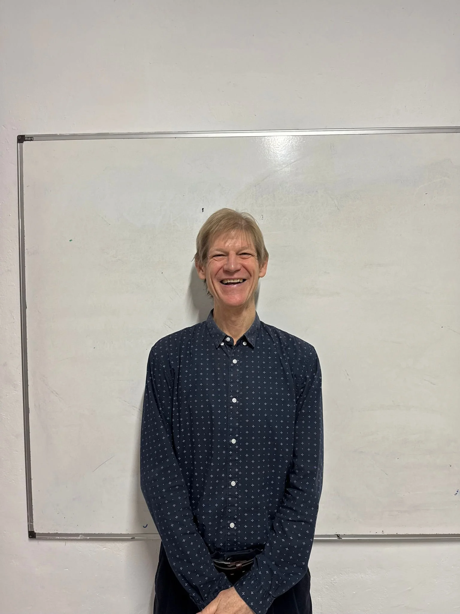 A man standing on a forest trail, smiling, wearing glasses, a flat cap, a black jacket, plaid shirt, blue jeans, and hiking boots.