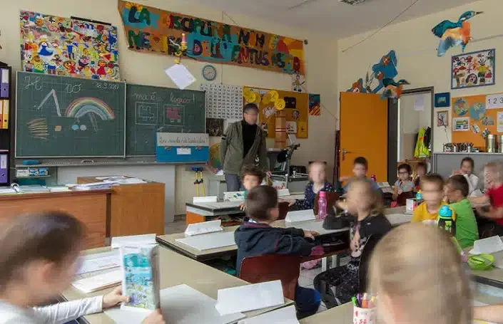 Classroom with children seated at desks, a teacher standing near the chalkboard, and colorful decorations including hanging paper crafts and a rainbow drawing.