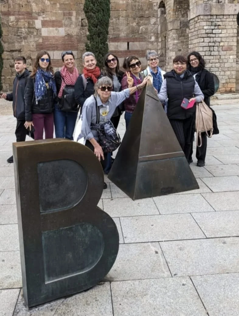 Group of ten women and one young man posing outdoors near a large black letter 'B' and former Roman temple ruins in the background.