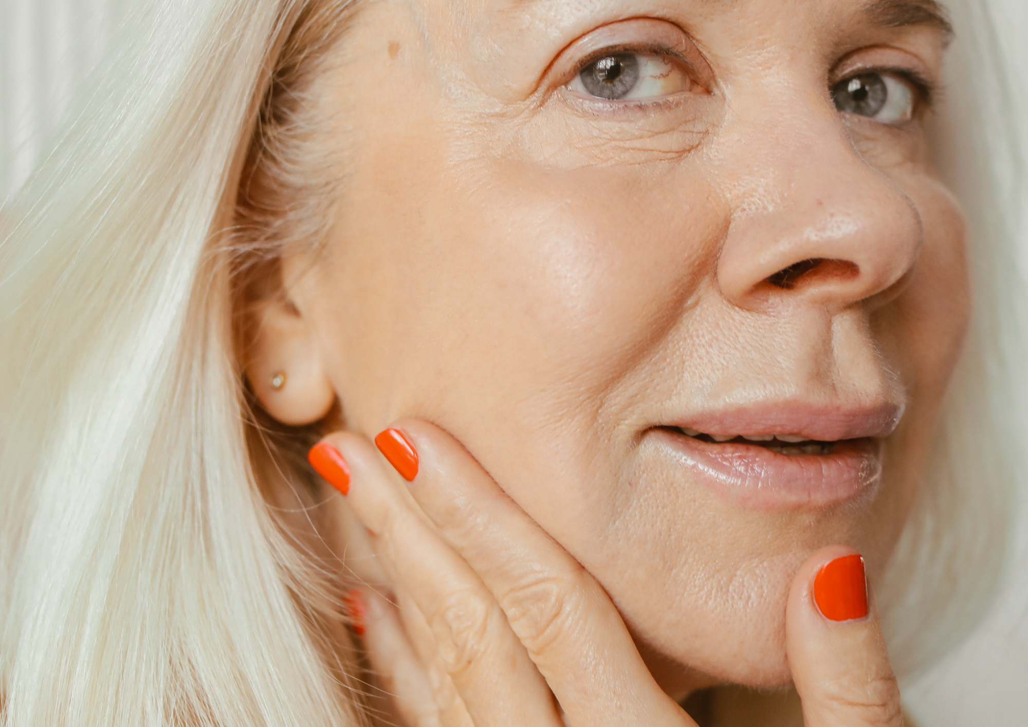 Close-up of a woman with blonde hair touching her face, showing clear skin, with visible eye, nose, and lips, and wearing orange nail polish.