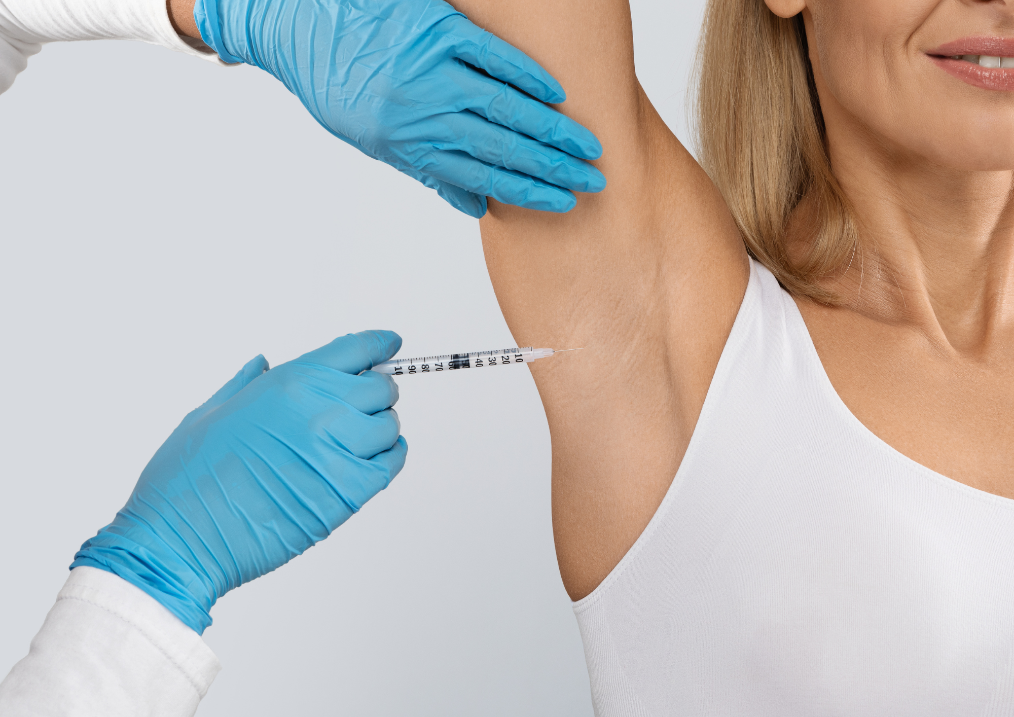 A woman in a white sleeveless top receives a vaccine shot in her upper arm from a healthcare professional wearing blue gloves.