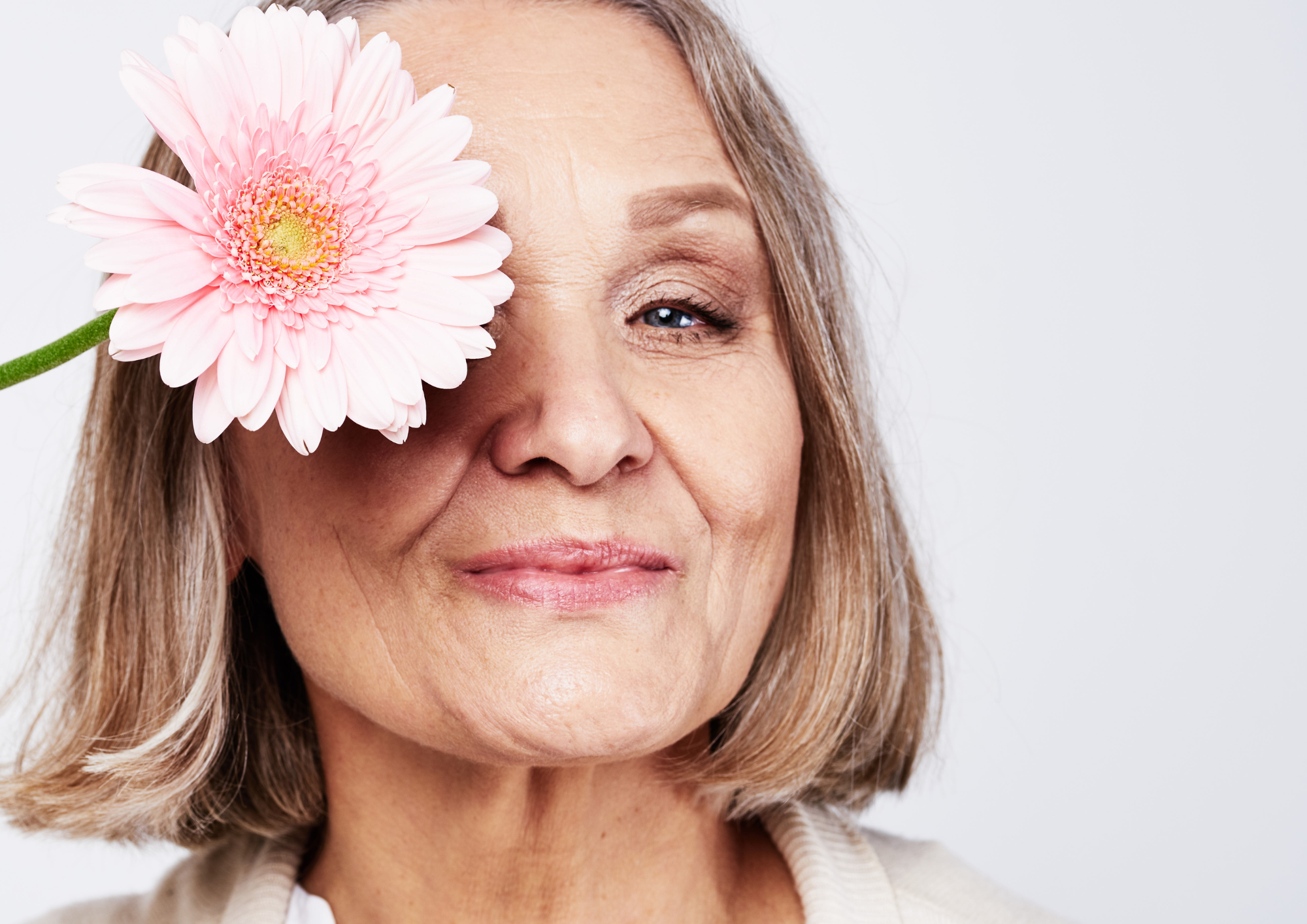 An elderly woman with blonde hair and light skin holding a pink daisy flower over her right eye, smiling gently against a plain white background.