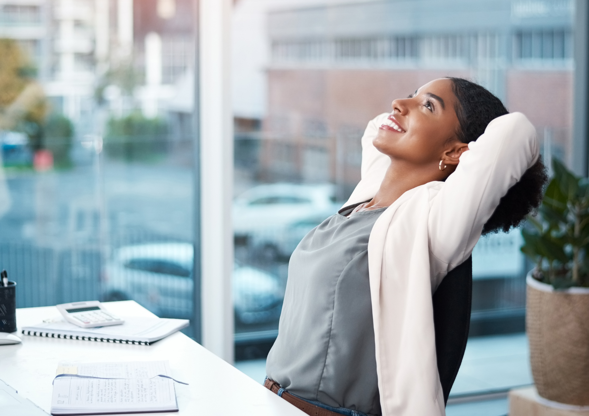 A woman relaxing at her office desk with her hands behind her head, smiling and looking upward.