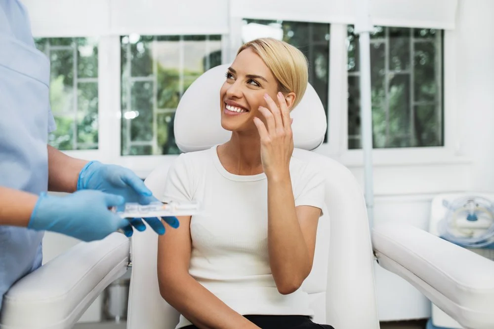 A woman smiling and sitting in a dental chair at a dental office, talking to a dental professional wearing gloves, who is handing her a tray of dental tools.