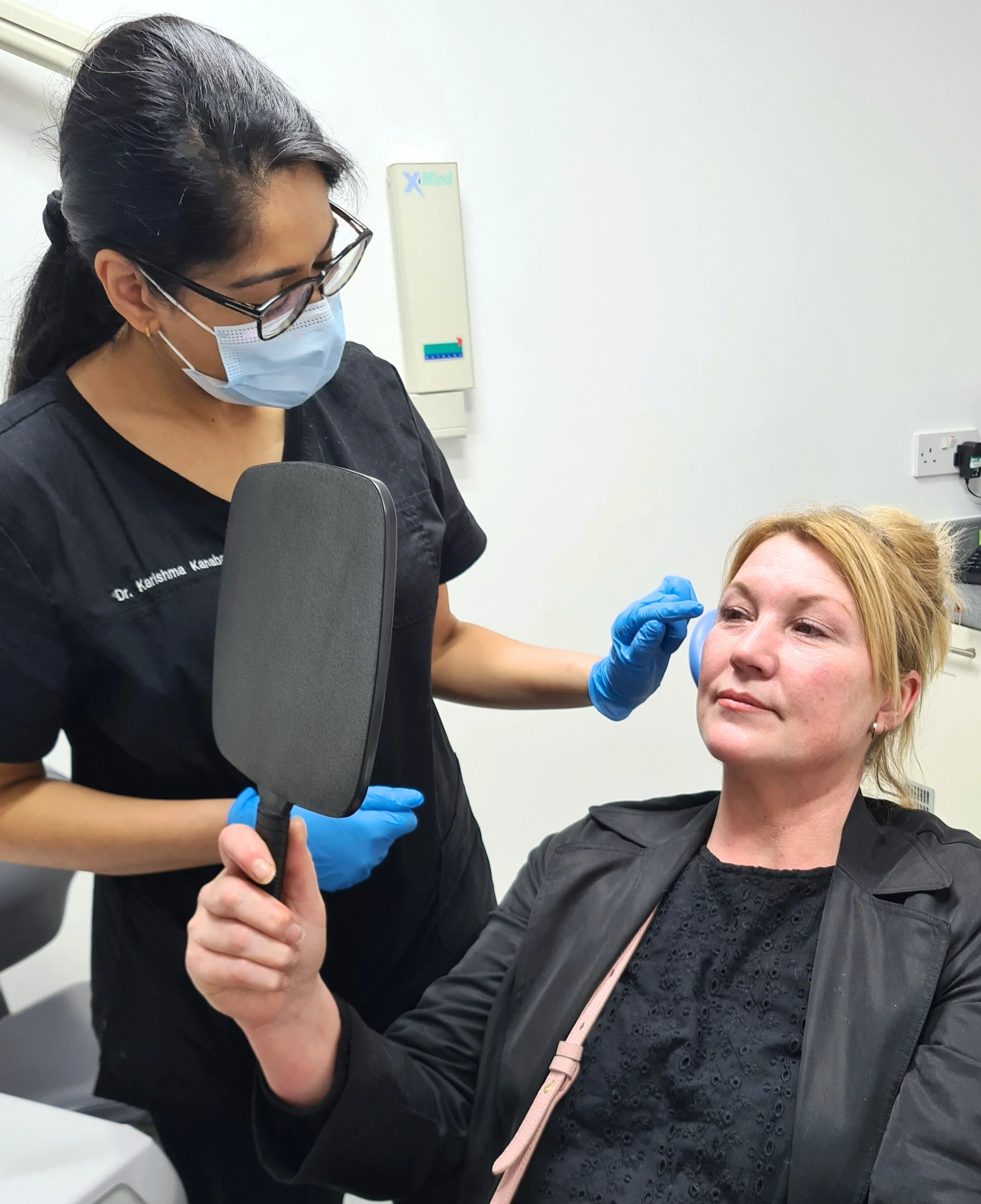 A woman with blonde hair and black clothing holding a mirror while a female healthcare professional examines her face. The healthcare professional is wearing a mask, glasses, a black uniform, and blue gloves.