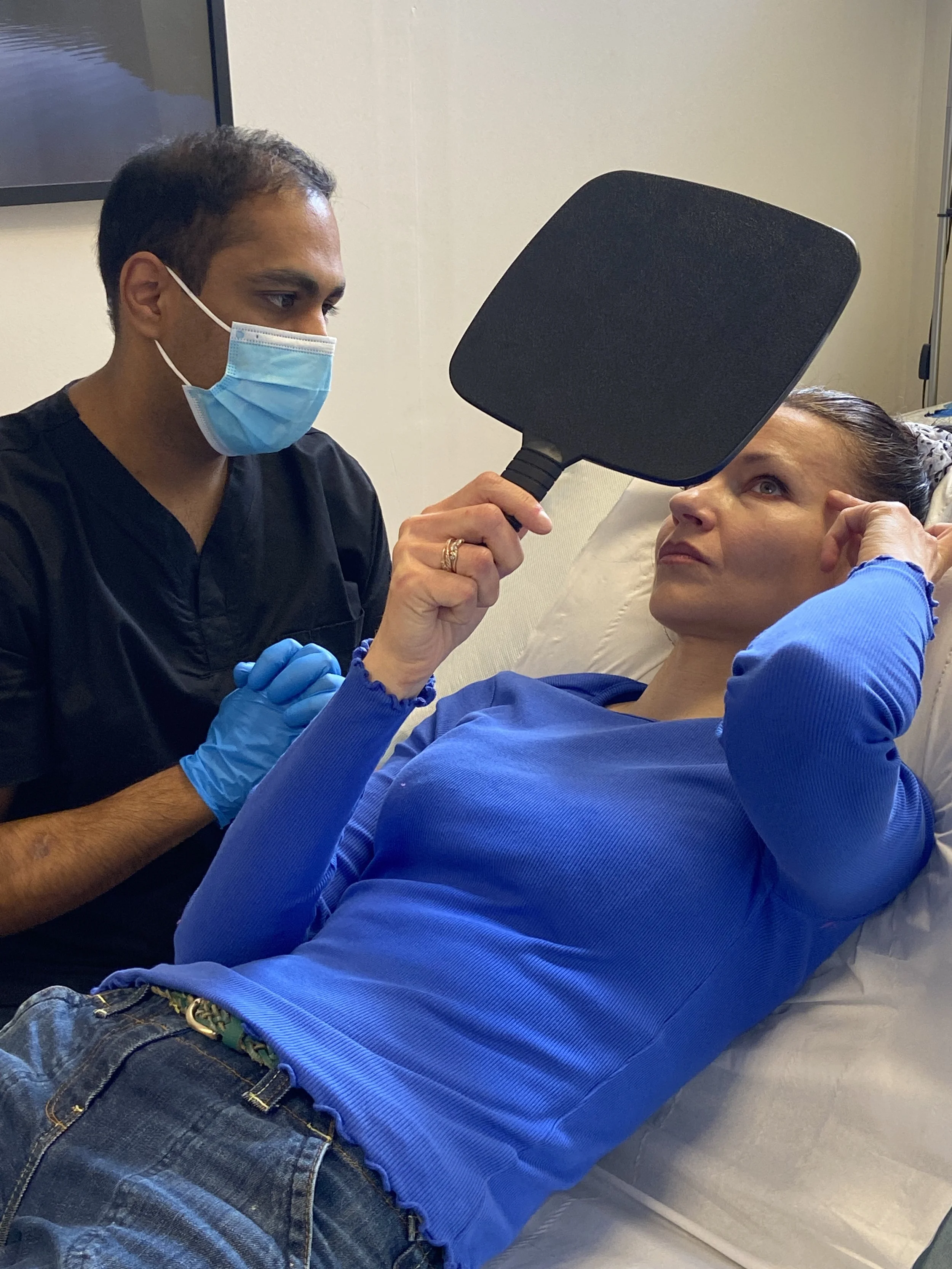 A healthcare professional wearing a face mask and gloves examines a patient lying in bed, holding a black mirror in front of her face.