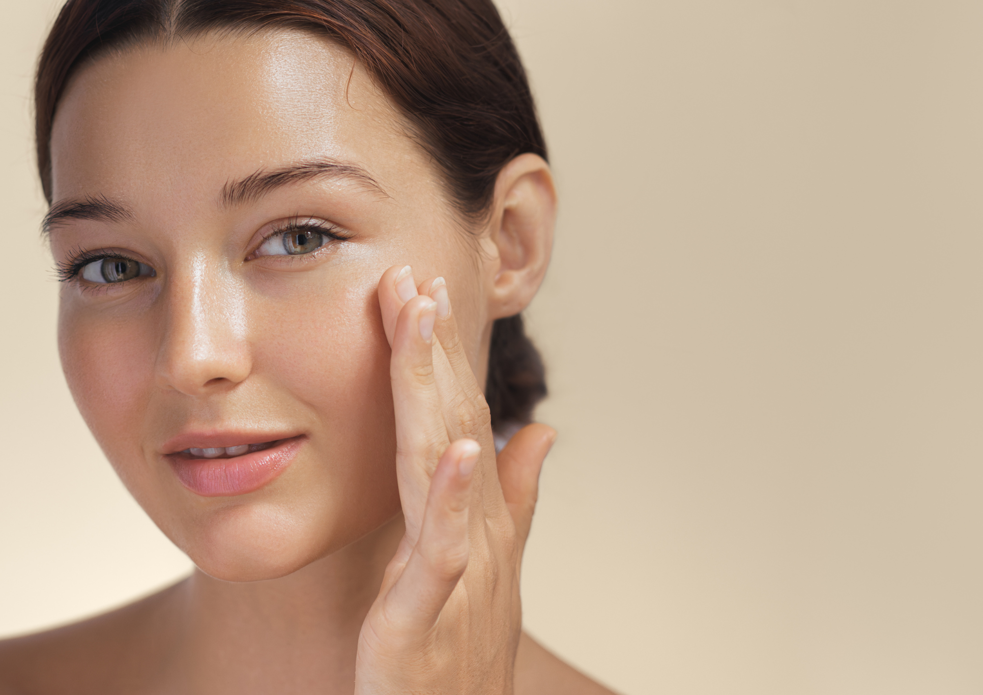 Close-up of a woman with fair skin and blue eyes applying moisturizer to her cheek, with a neutral beige background.