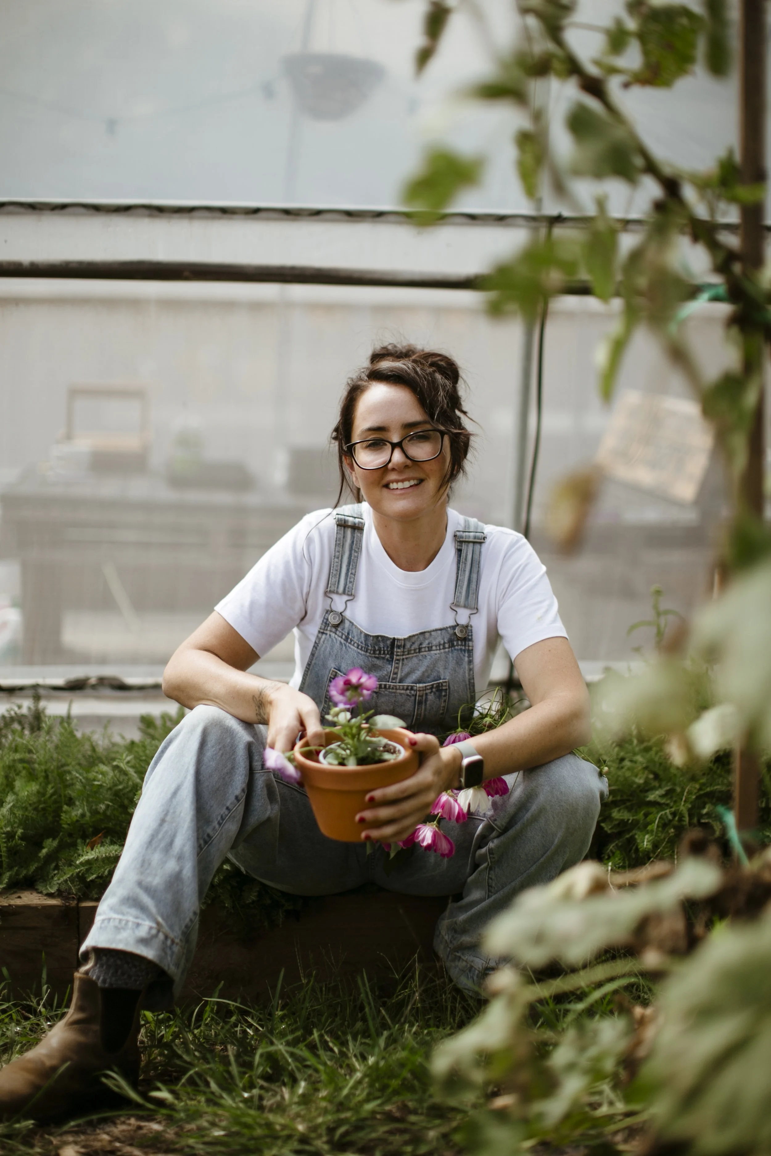 “Woman sitting in a flower garden holding a small potted plant, wearing denim overalls, surrounded by soft natural greenery in a calm, slow living farm setting.”