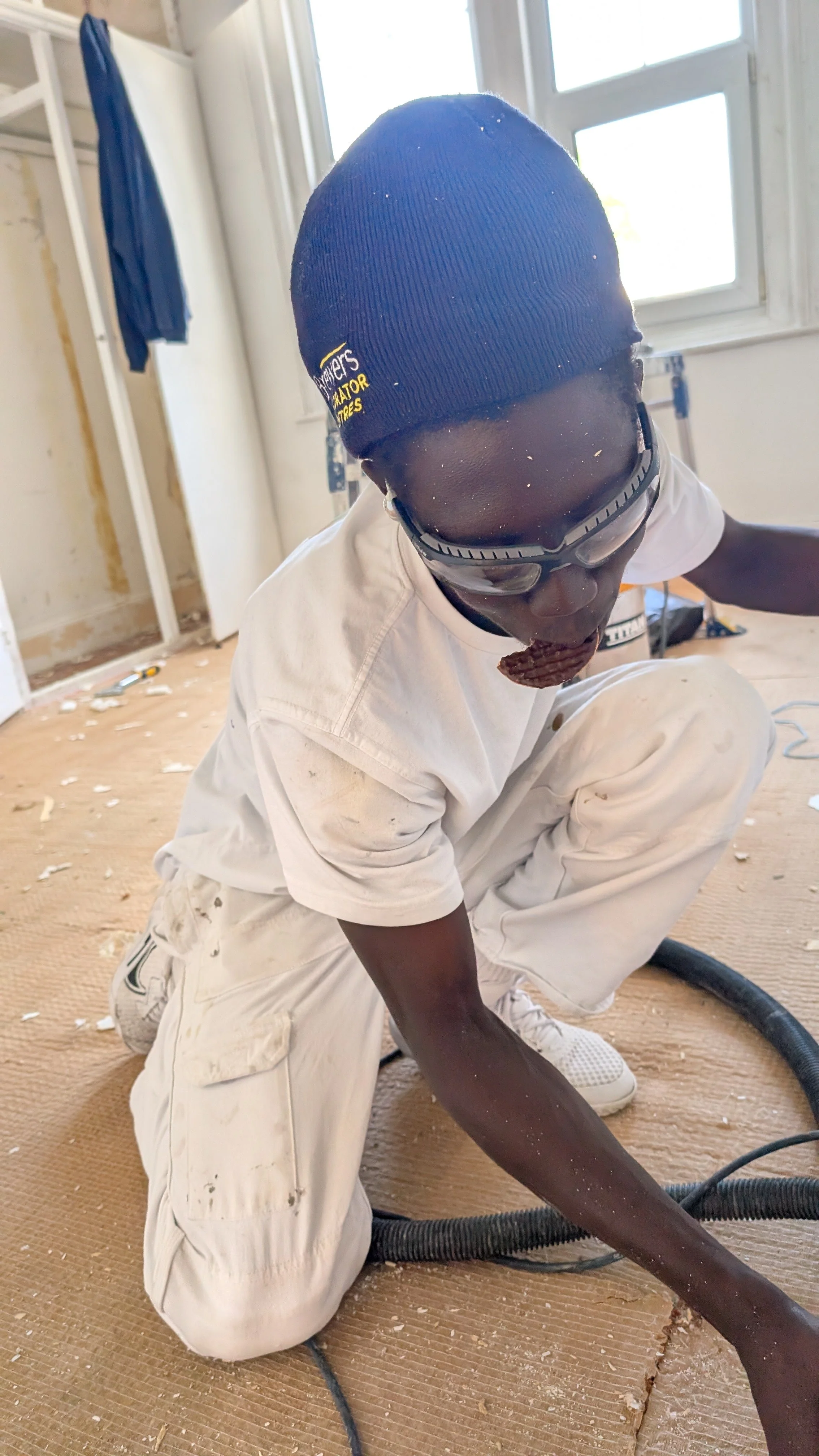 A person kneeling on the floor in a room under construction or renovation, wearing a black beanie, goggles, white shirt, and white pants, surrounded by construction debris.